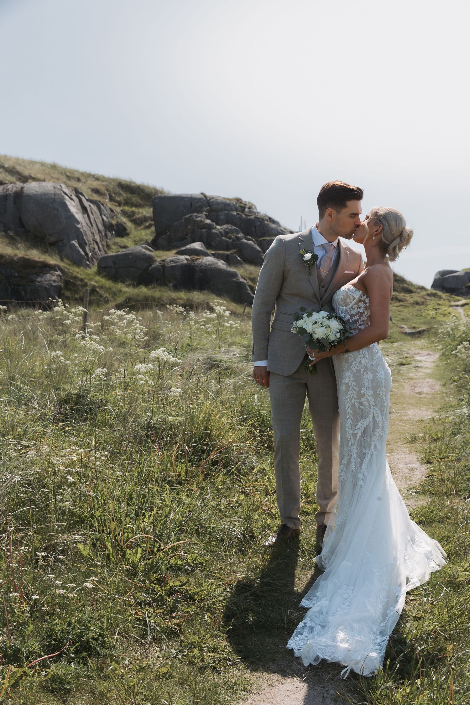 Bride and groom kiss on scenic rocky hillside path, she holds white bouquet in lace wedding gown.