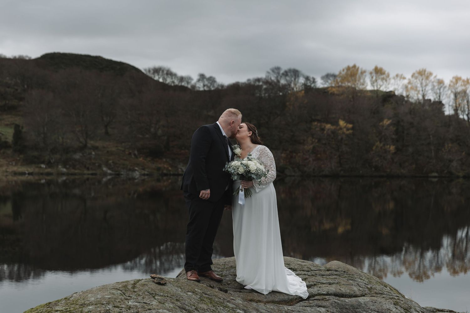 Bride and groom share a kiss on a lakeside rock in the Lake District, surrounded by autumn hills and calm water.