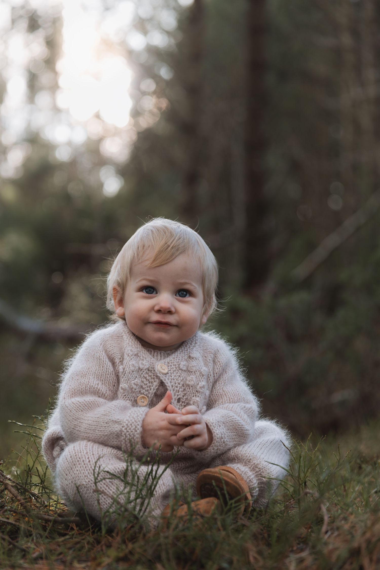 A small child wearing a cozy beige sweater sits on grass in a forest setting during golden hour.