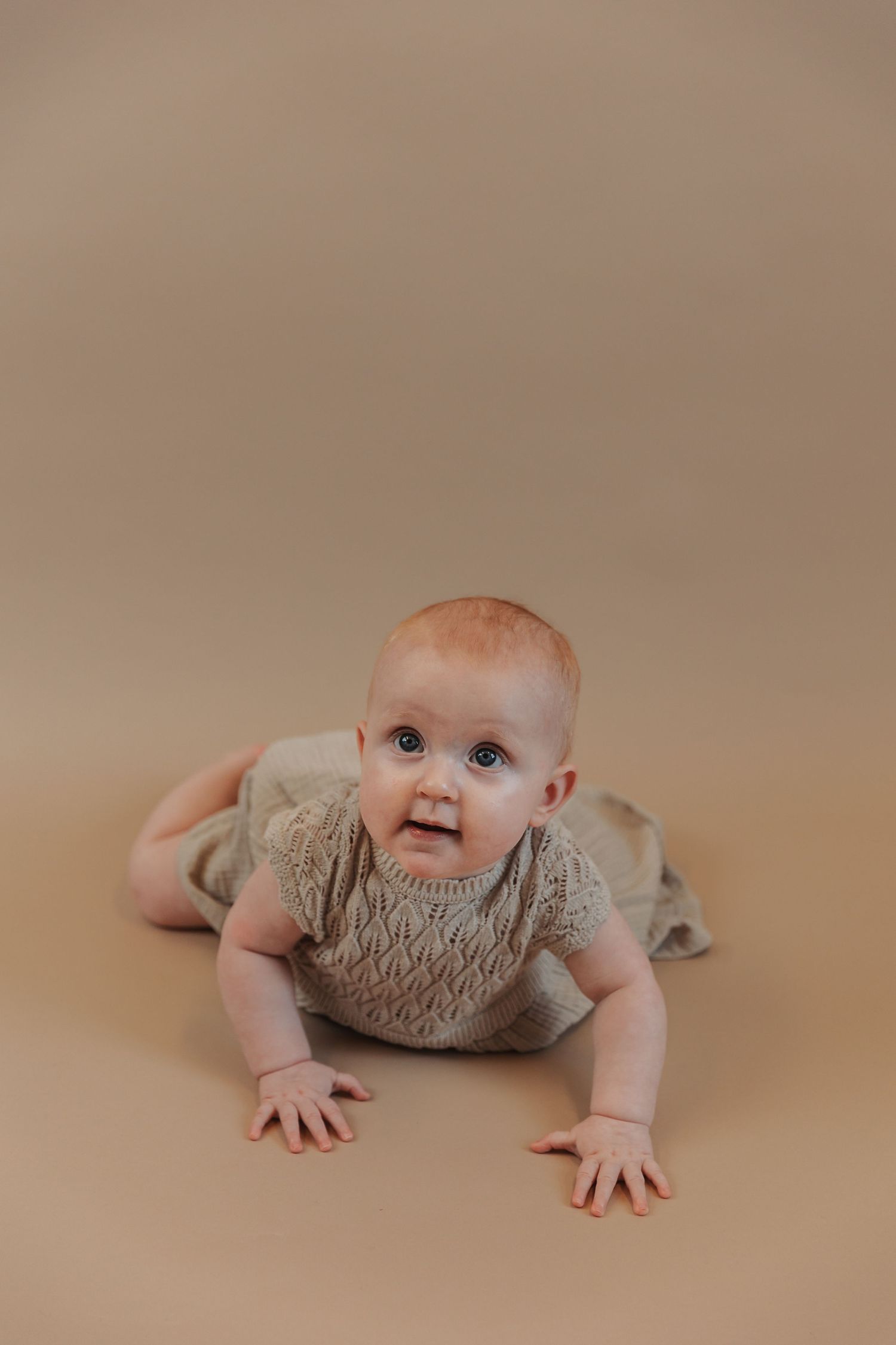 An infant in a beige smocked romper lies on their belly against a neutral backdrop.