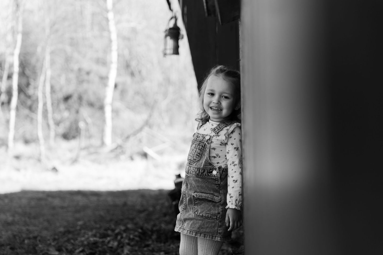 A black and white image shows a child standing by a doorway, wearing a floral top and smiling.