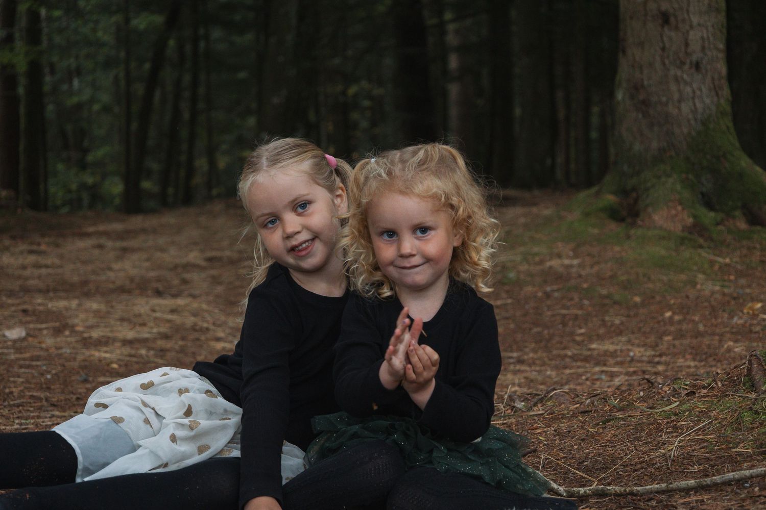Two children in dark clothing sitting together on forest ground.