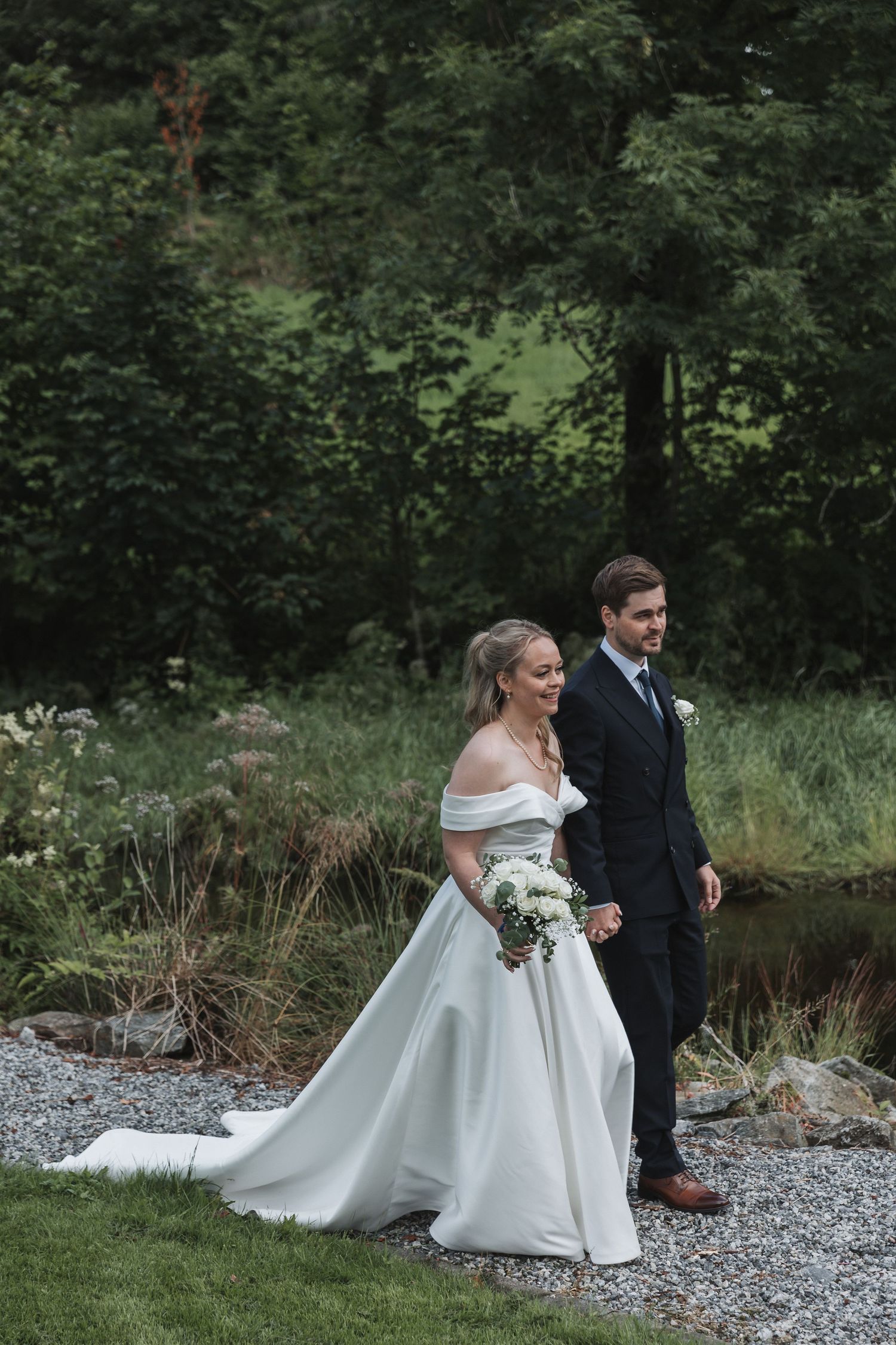 Bride in off-shoulder gown and groom walking hand in hand on gravel path in lush garden.