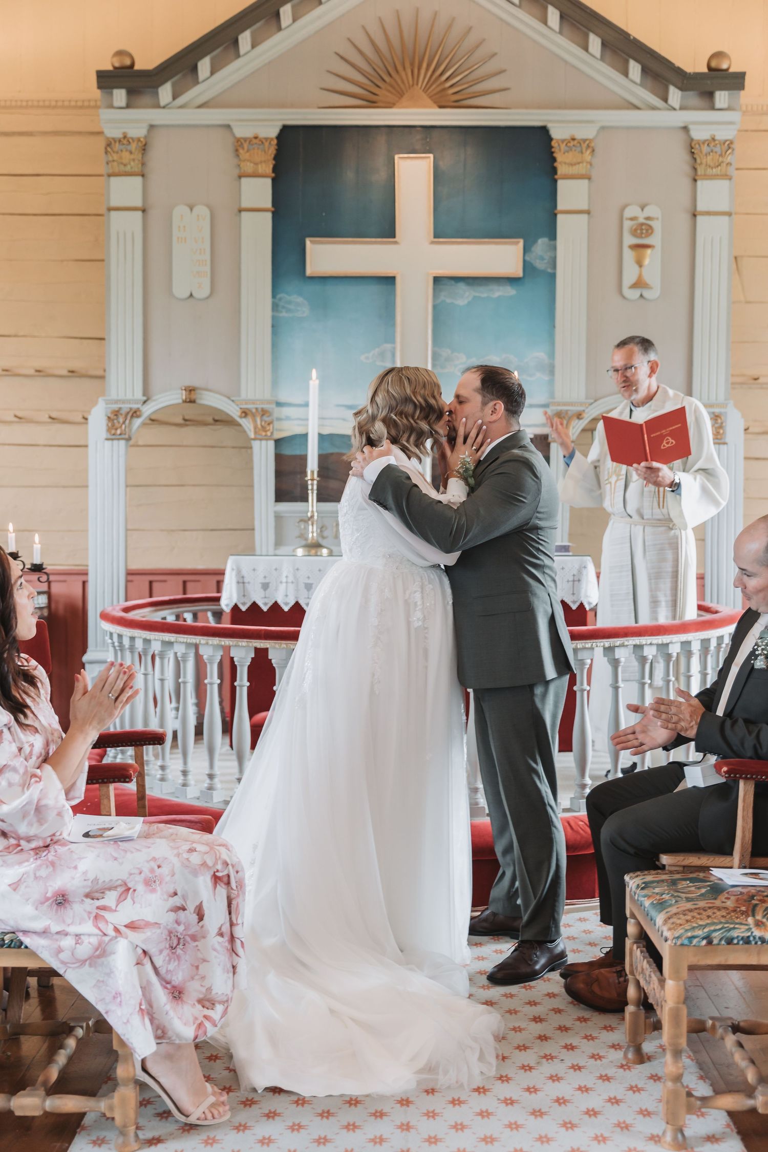 Bride and groom share first kiss at church altar during intimate wedding ceremony with officiant and guests applauding.