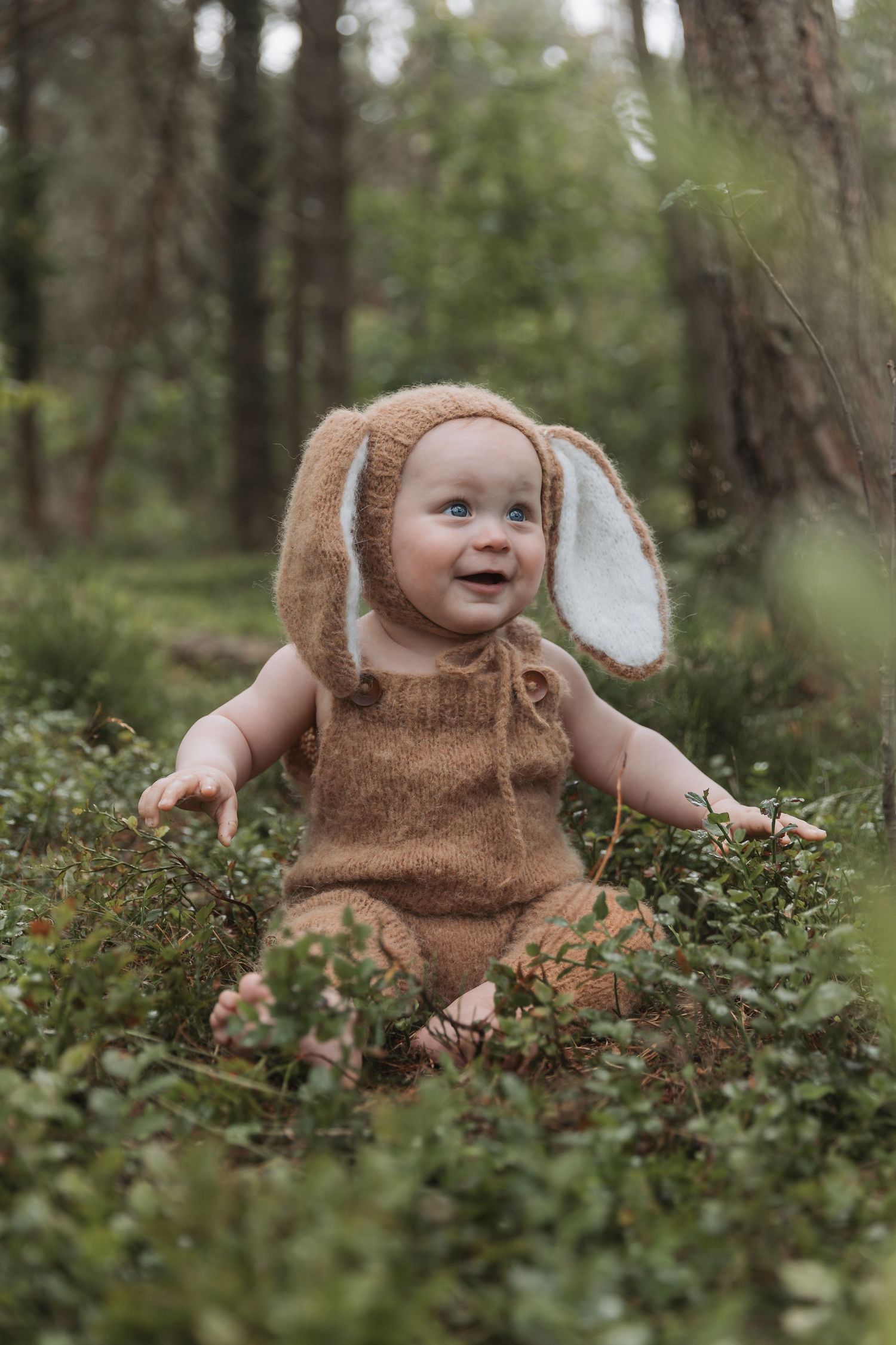 A baby wearing a brown bunny costume sits happily among green foliage in a forest setting.