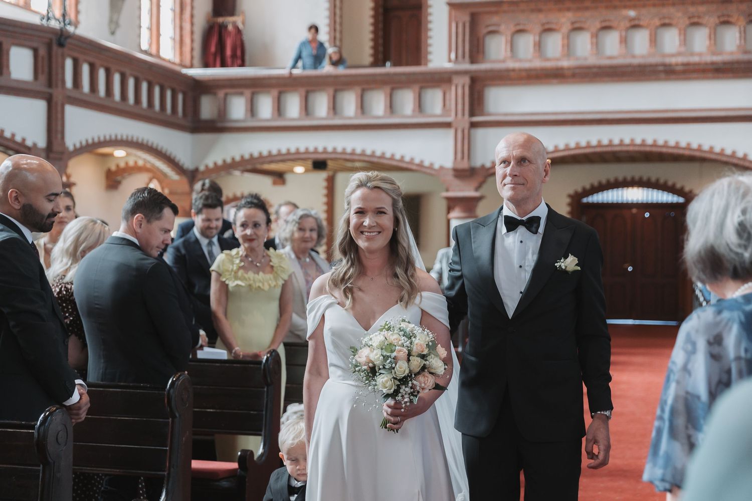 Bride walks down church aisle with escort, holding bouquet, guests watching in elegant wood-paneled wedding ceremony.