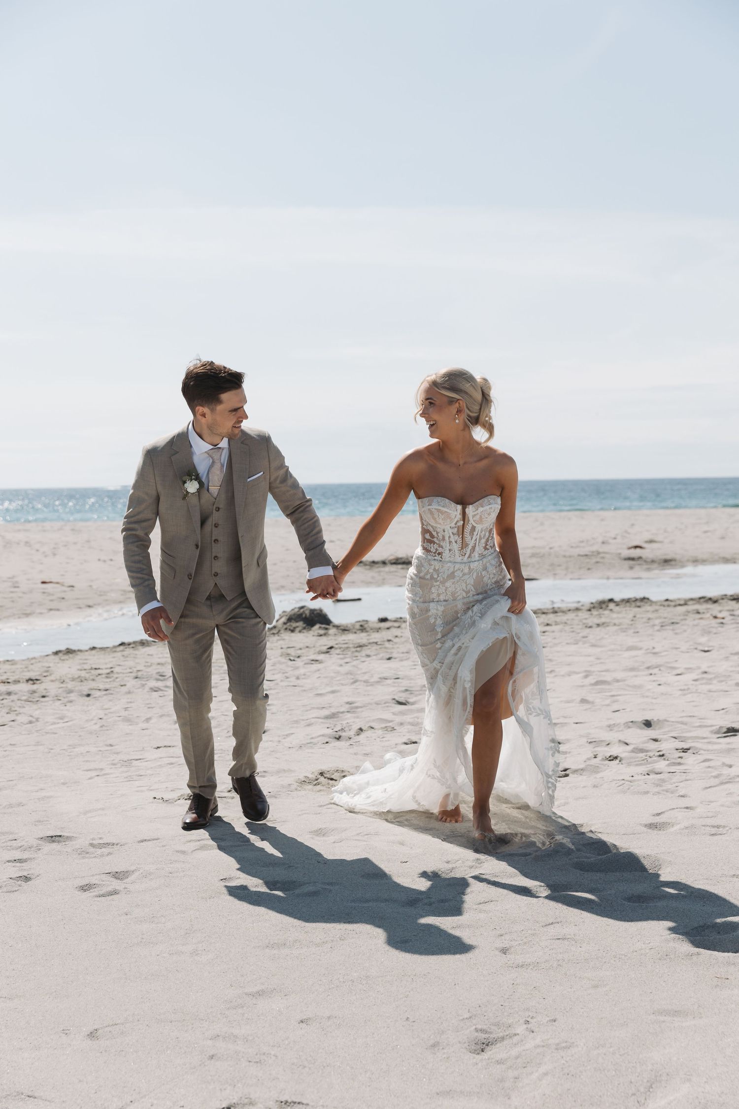 A couple in wedding attire playfully runs and dances together on a scenic beach with mountains in the background.