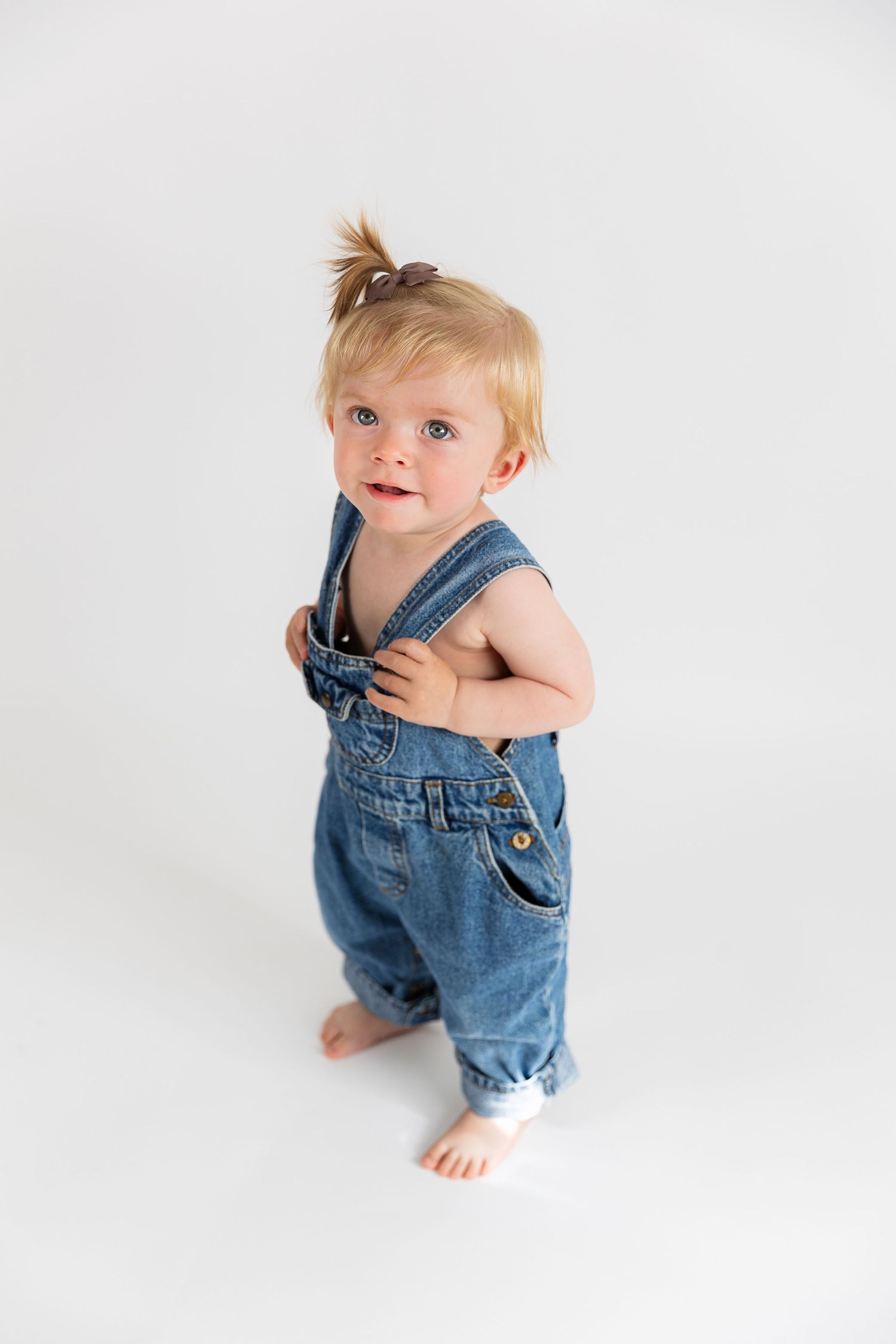 Adorable toddler in denim overalls sits and stands in three poses against white backdrop in professional photo series.