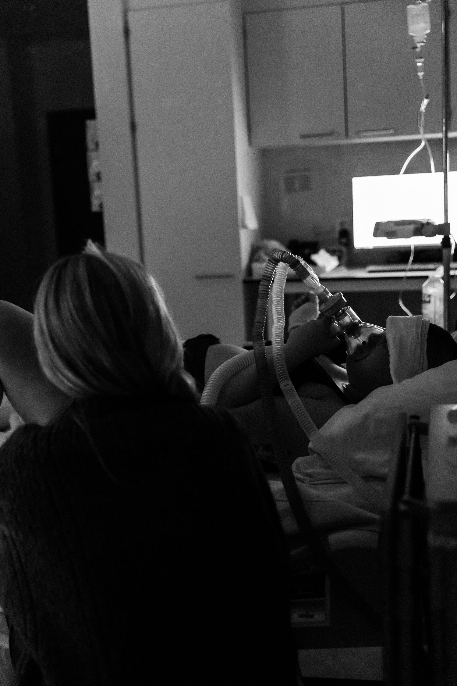 Dark monochrome photo of someone lying in a hospital room with medical equipment visible in the background.