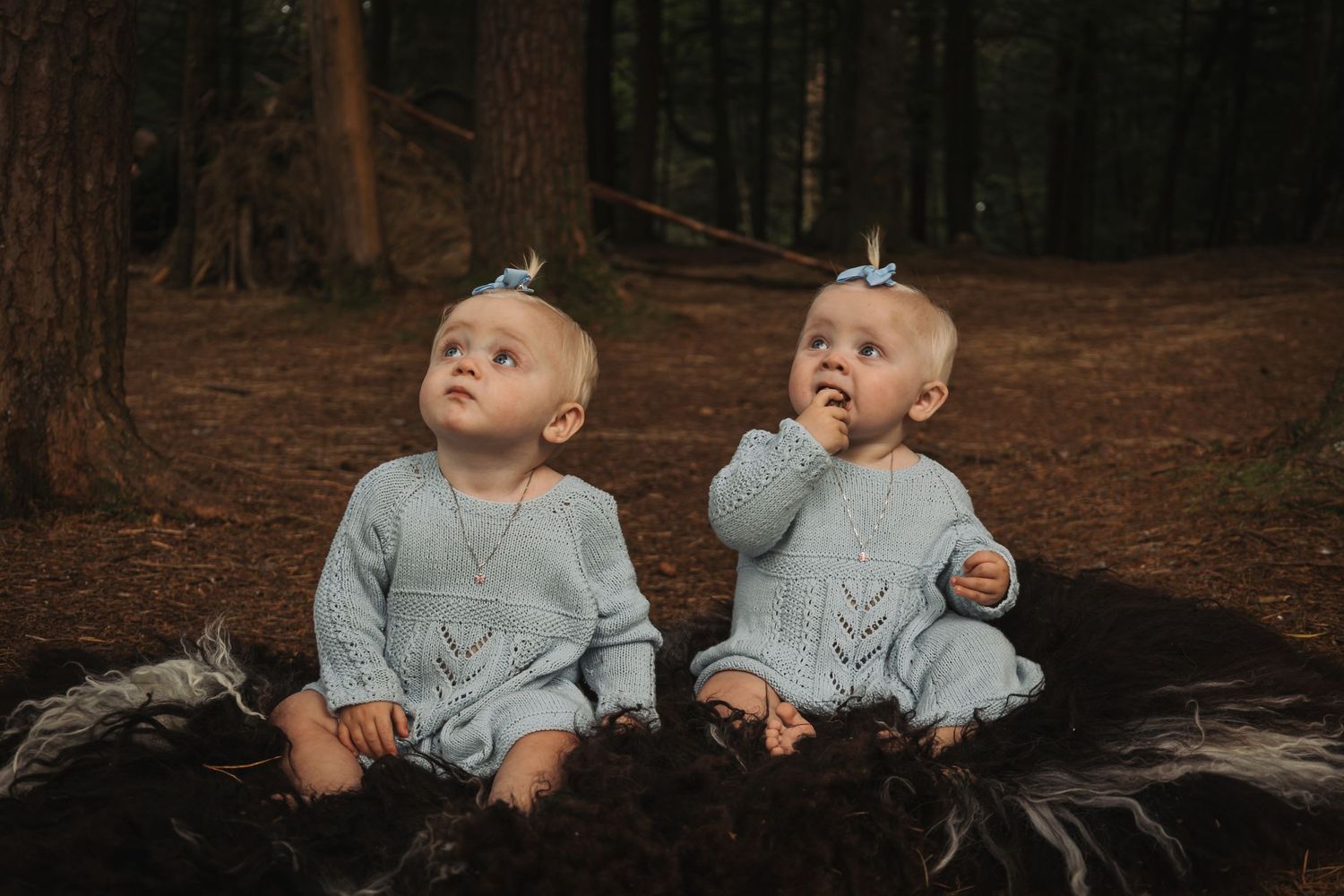 Two babies in matching gray outfits sit together on a dark blanket in the woods.