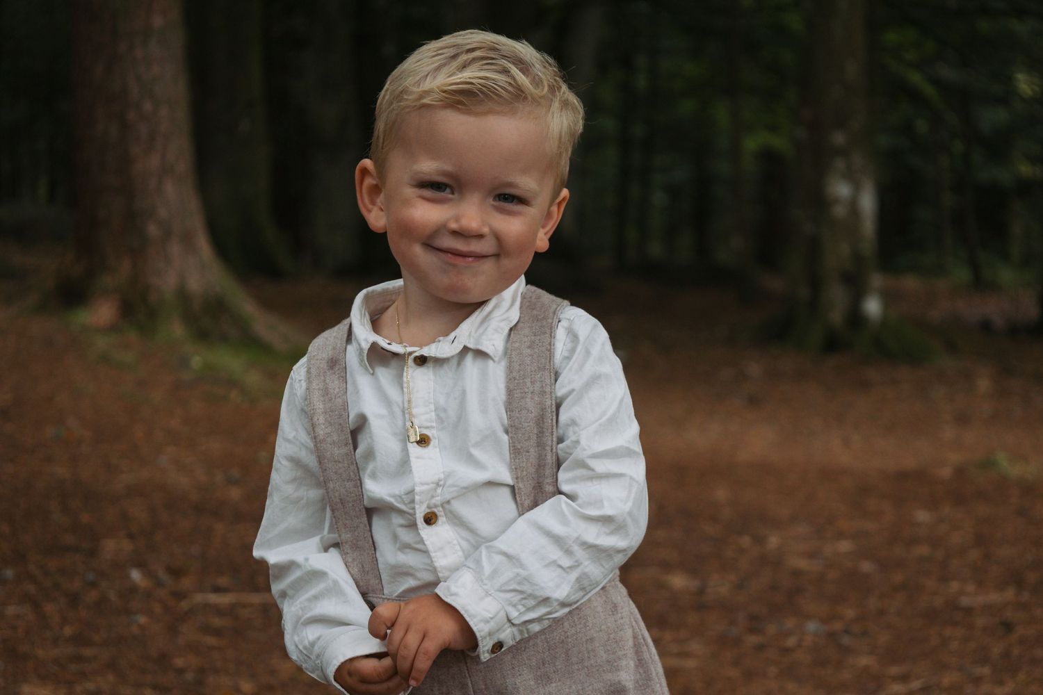 Smiling child in white button-down shirt and gray pants stands in forest setting.