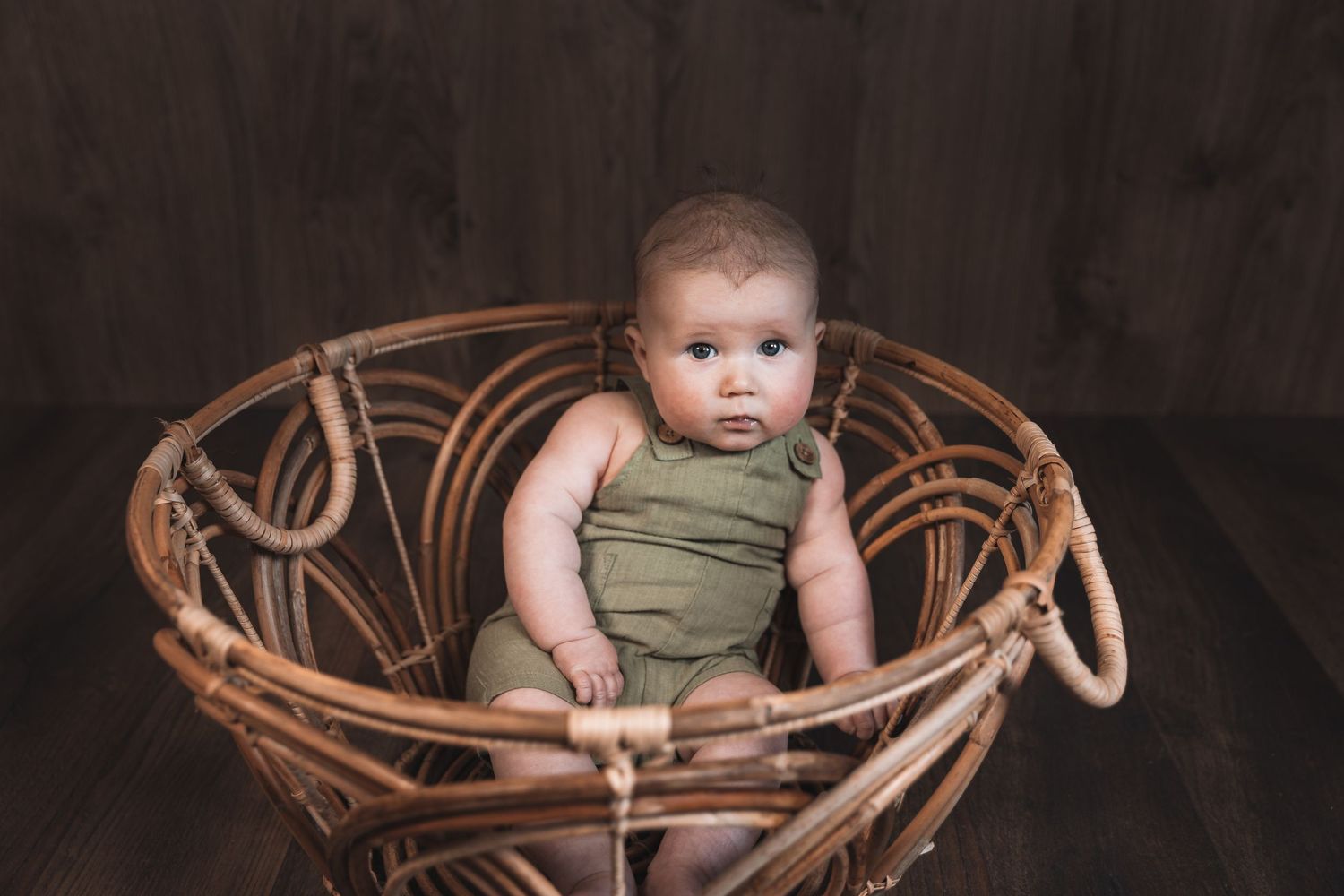Baby wearing olive green romper sits in vintage woven rattan peacock chair against dark backdrop.