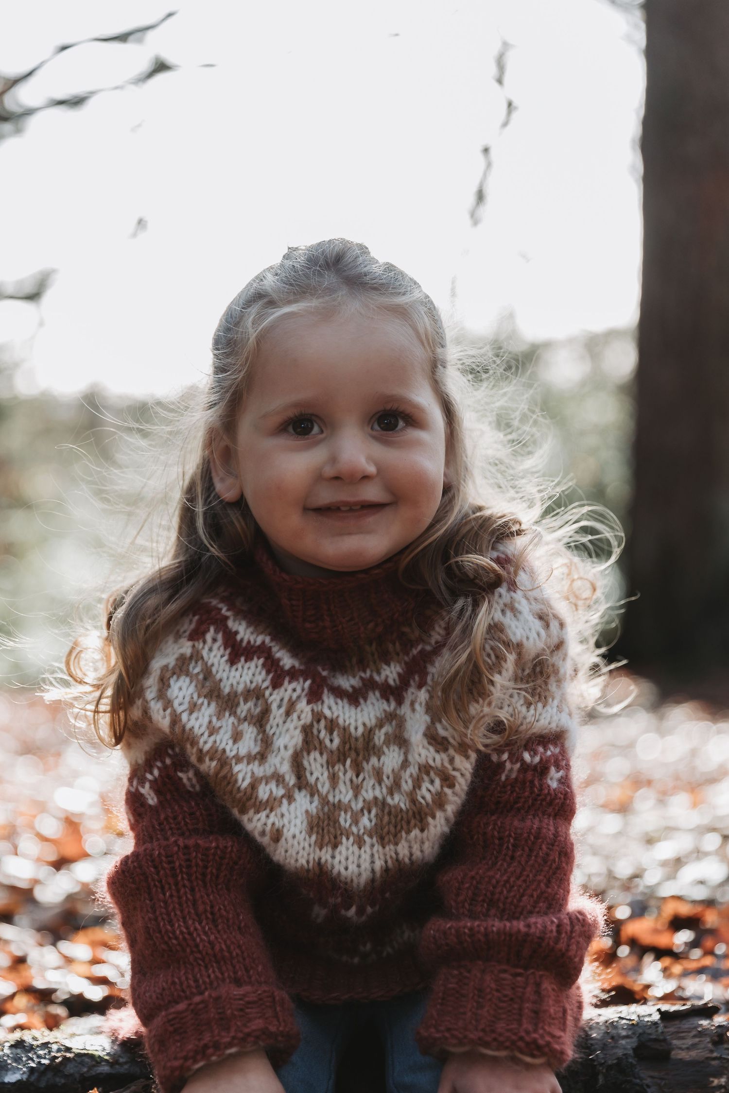 Small child wearing a brown and white knit sweater sits among autumn leaves on a crisp fall day.