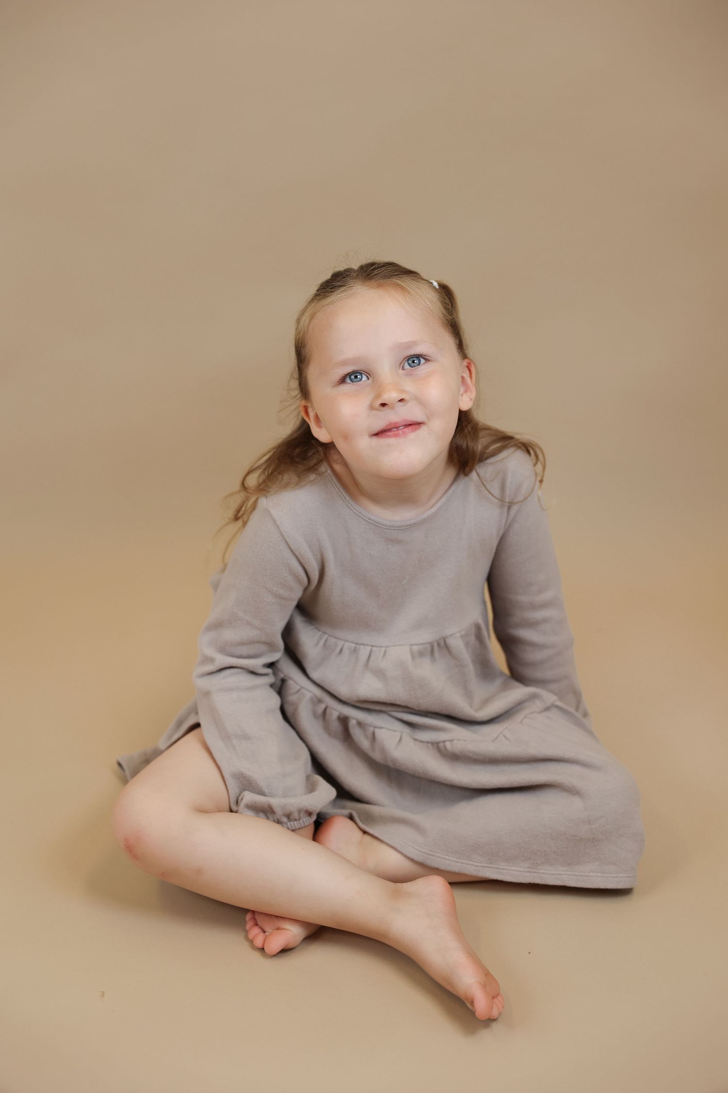 A young girl with blue eyes sits cross-legged in a gray long-sleeve dress on a beige background, smiling warmly.