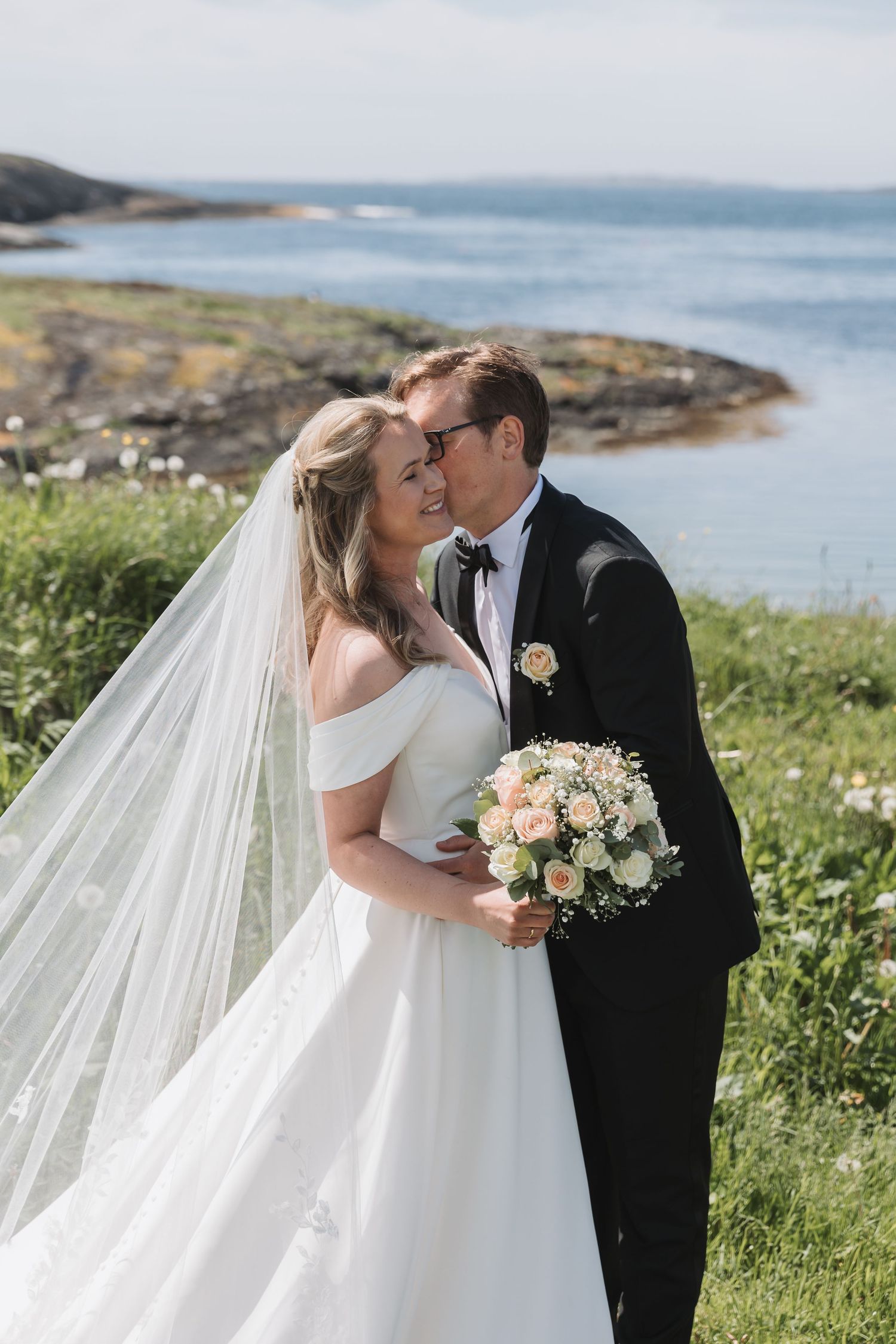 Bride and groom share a romantic kiss outdoors by the sea on their wedding day.