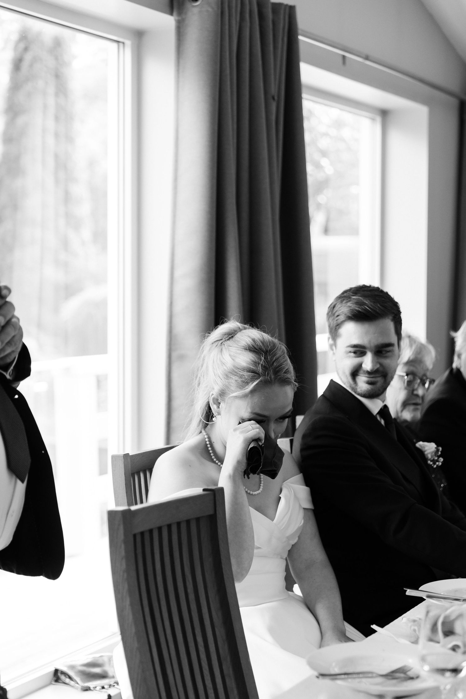 Black and white photo of emotional bride wiping tears while groom smiles warmly during wedding reception speeches.