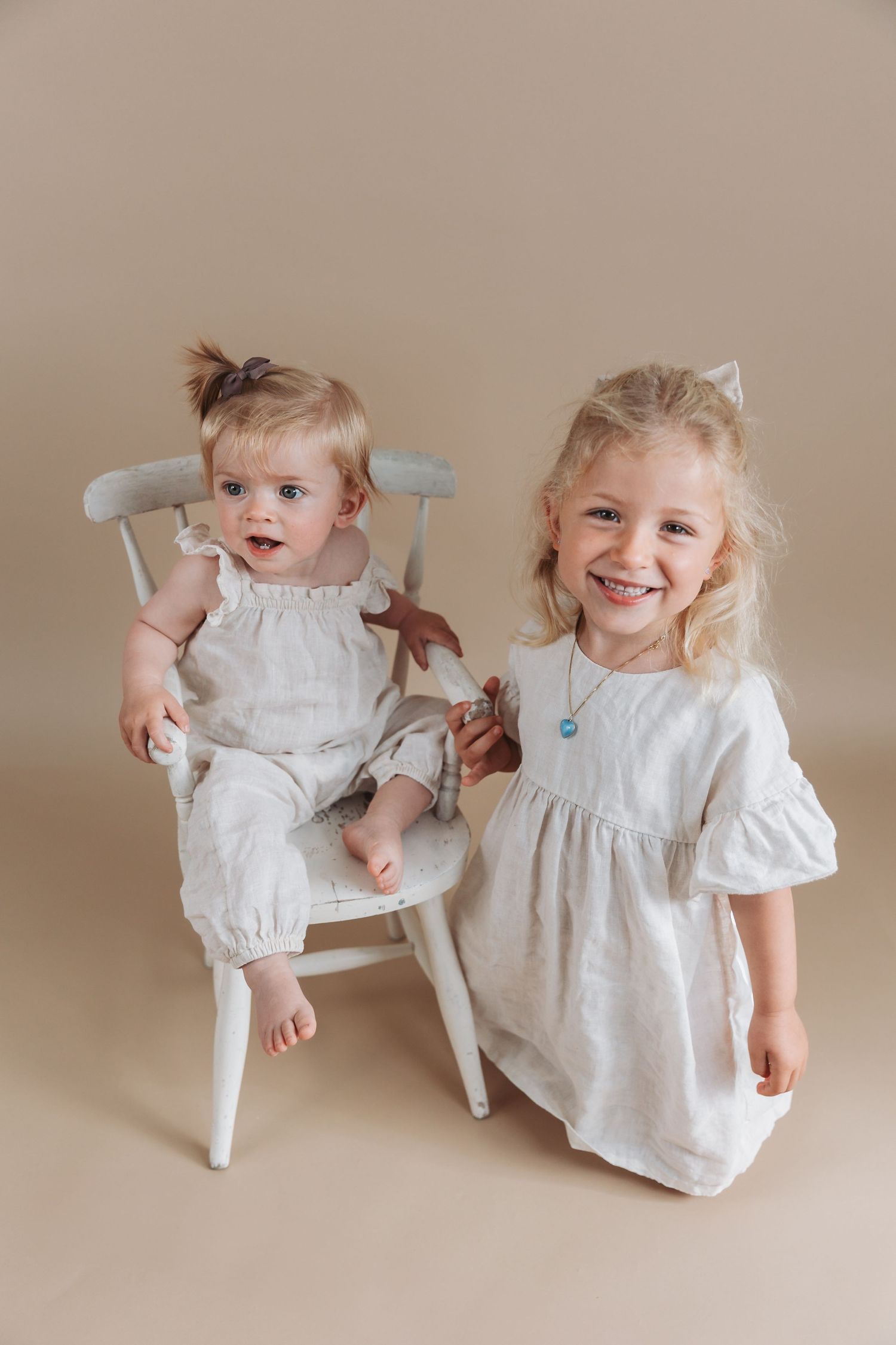 Two young children in white dresses pose together against a neutral backdrop, one seated in a white wooden chair.