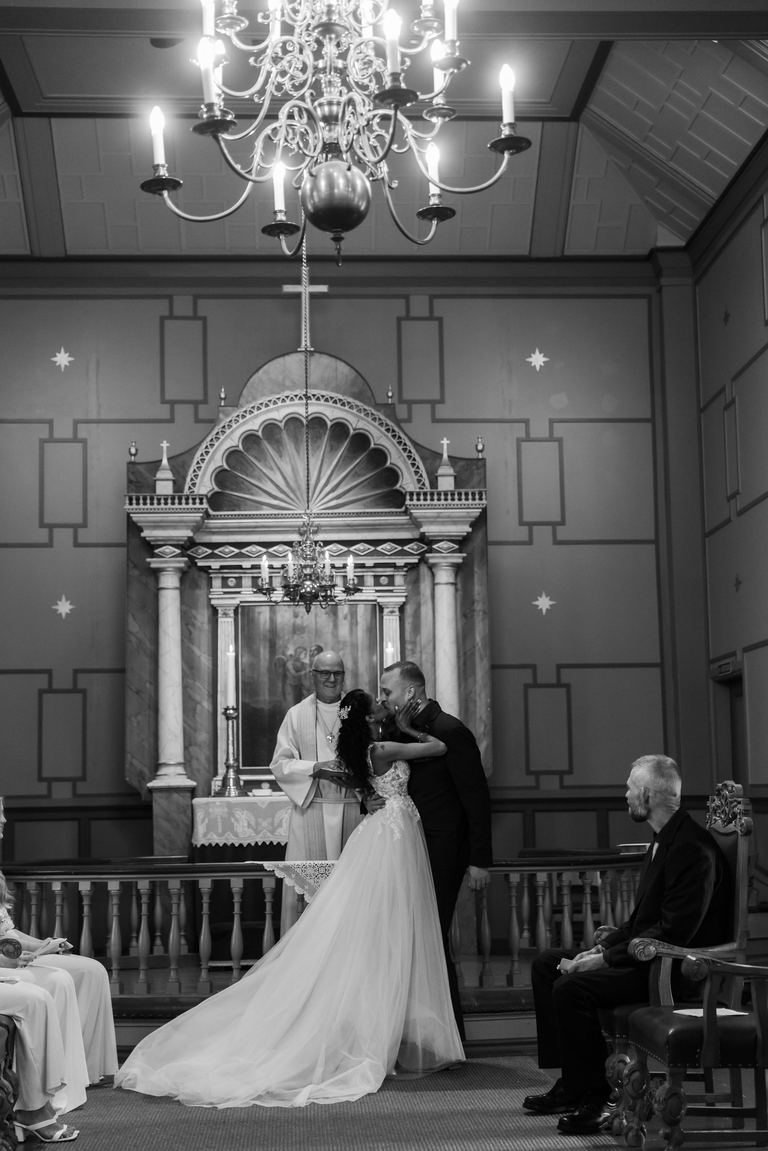 Black and white wedding ceremony photo of bride and groom at church altar with chandelier and officiant.