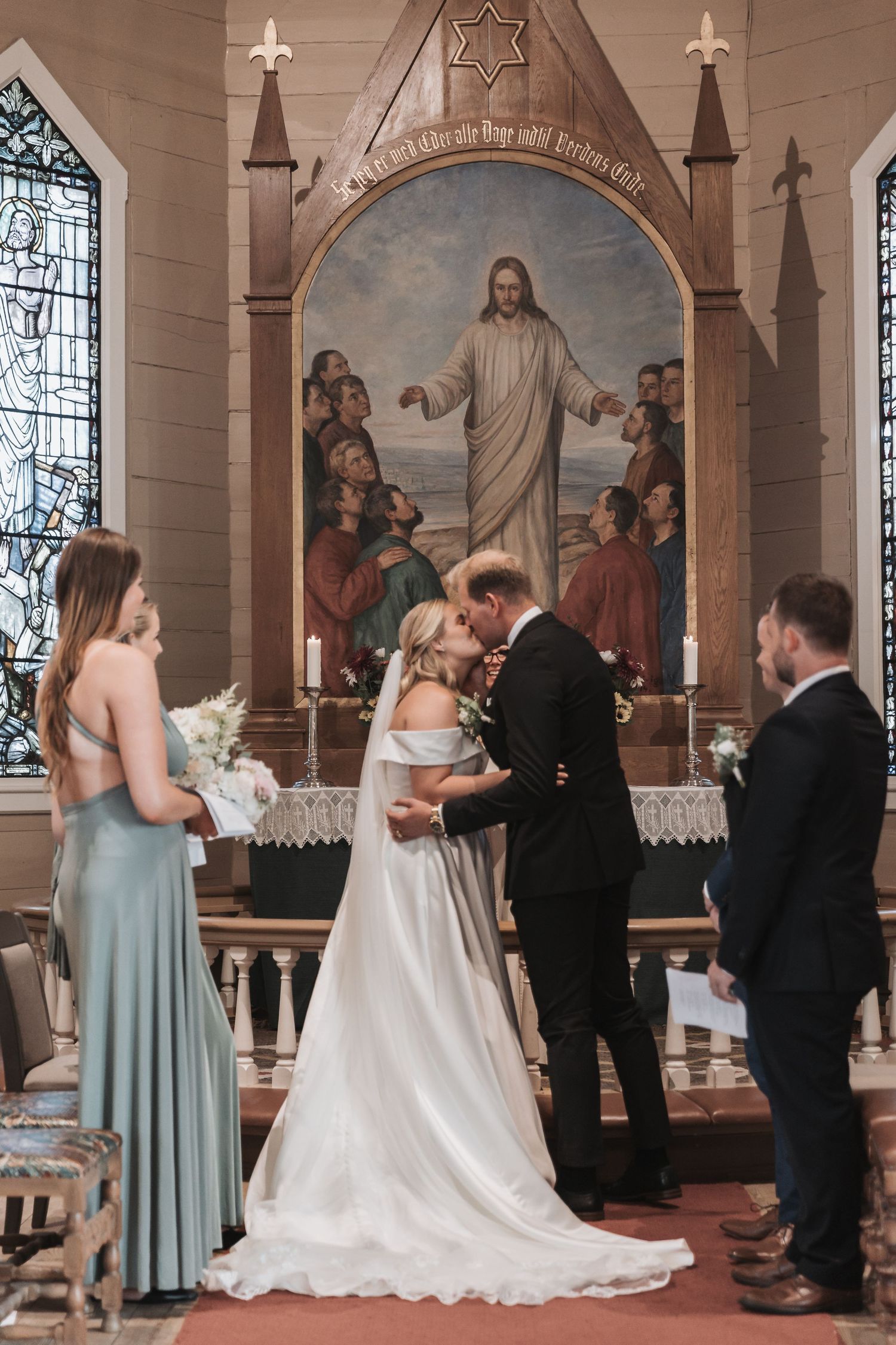 Bride and groom share first kiss at church altar with Jesus painting backdrop, flanked by bridesmaid and groomsman.