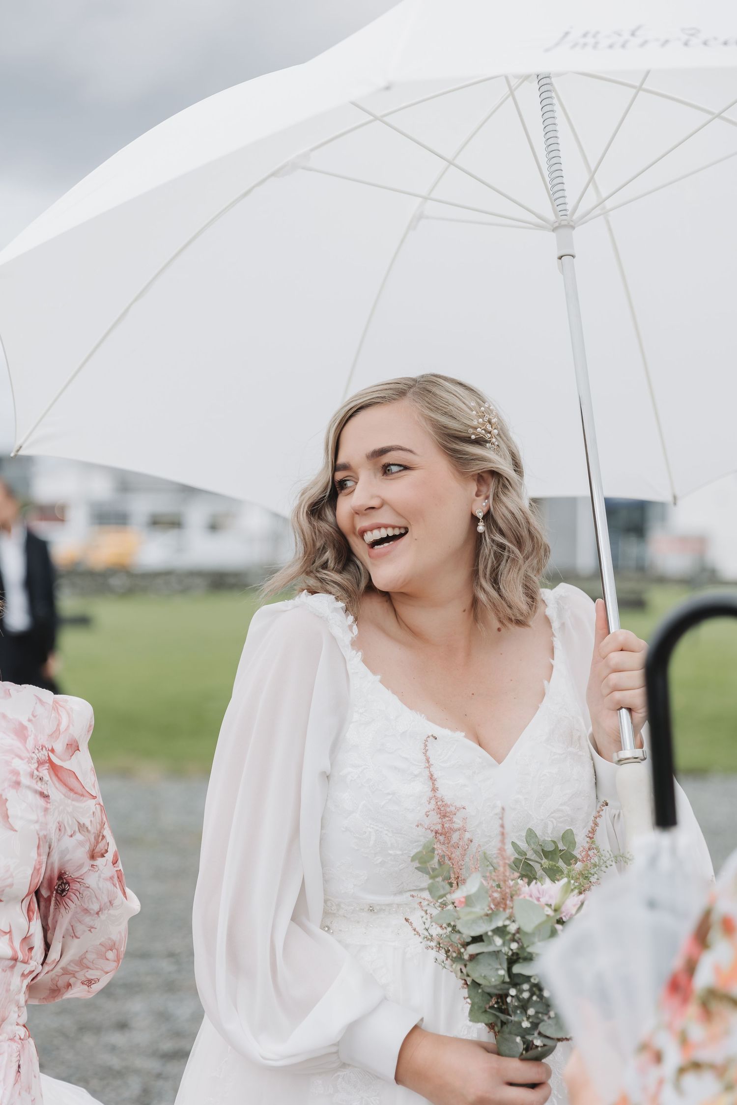 Smiling bride in white lace gown holding bouquet and white umbrella on a cloudy wedding day.