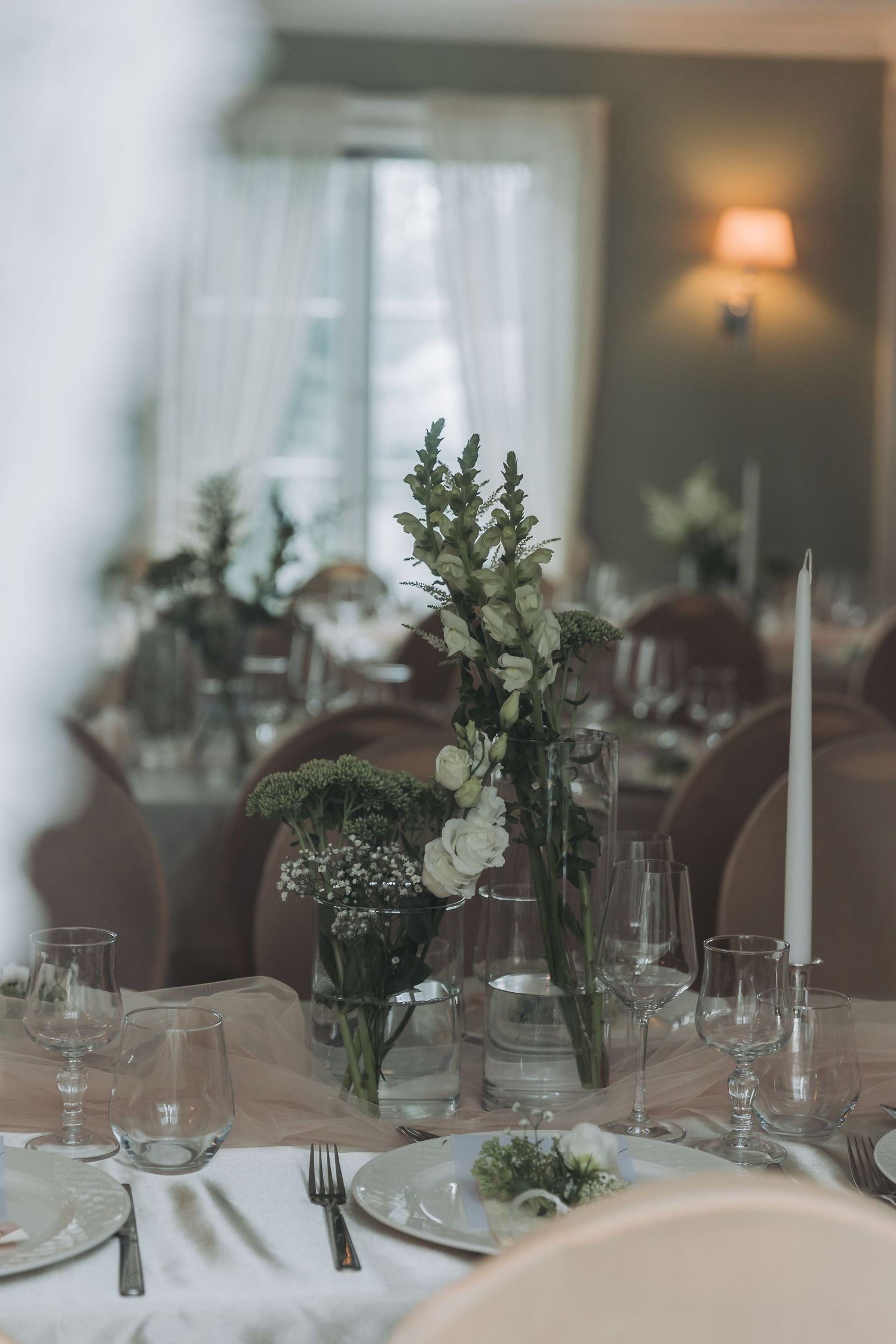 Elegant wedding reception table setting with white floral centerpieces, candles, and glassware in a softly lit venue.