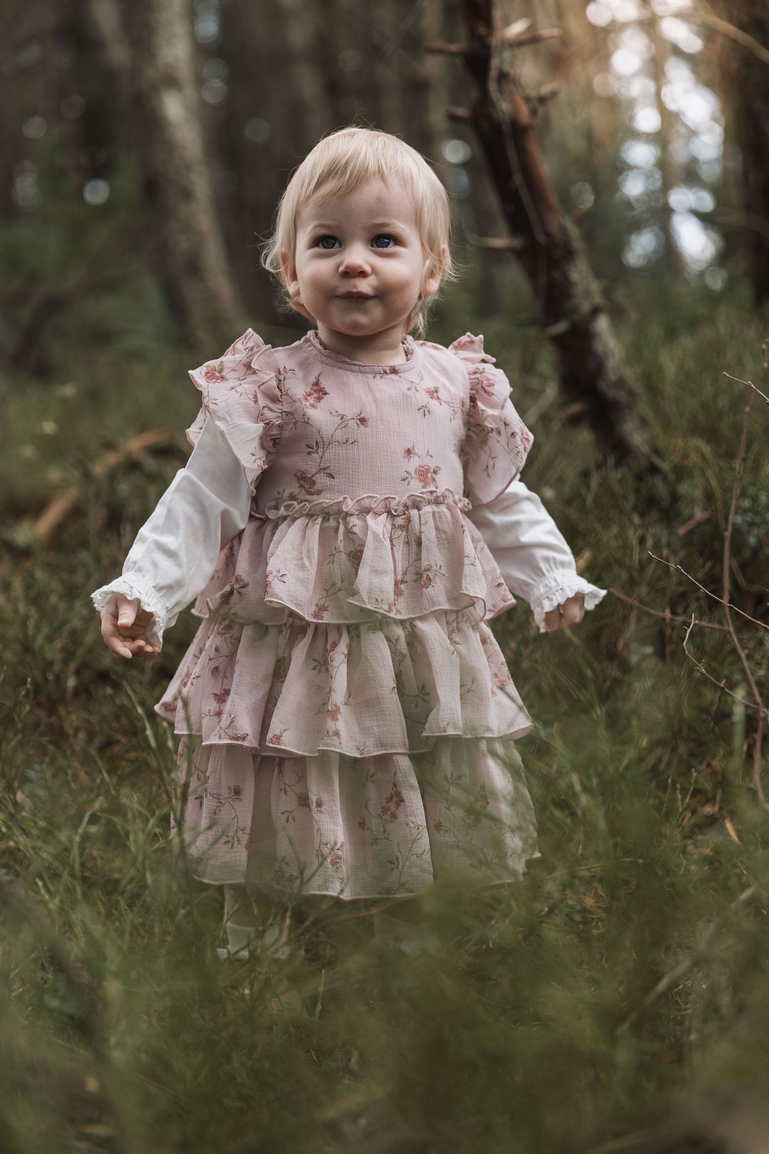 A toddler girl in a pink floral tiered ruffle dress stands outdoors in a lush green forest setting.