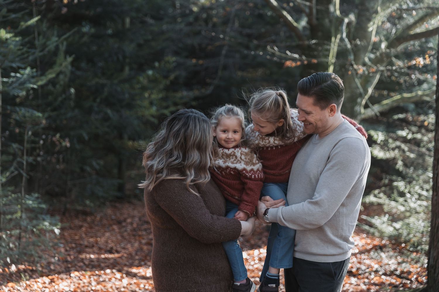 A family shares a tender moment together in an autumn forest setting with fallen leaves and evergreen trees.