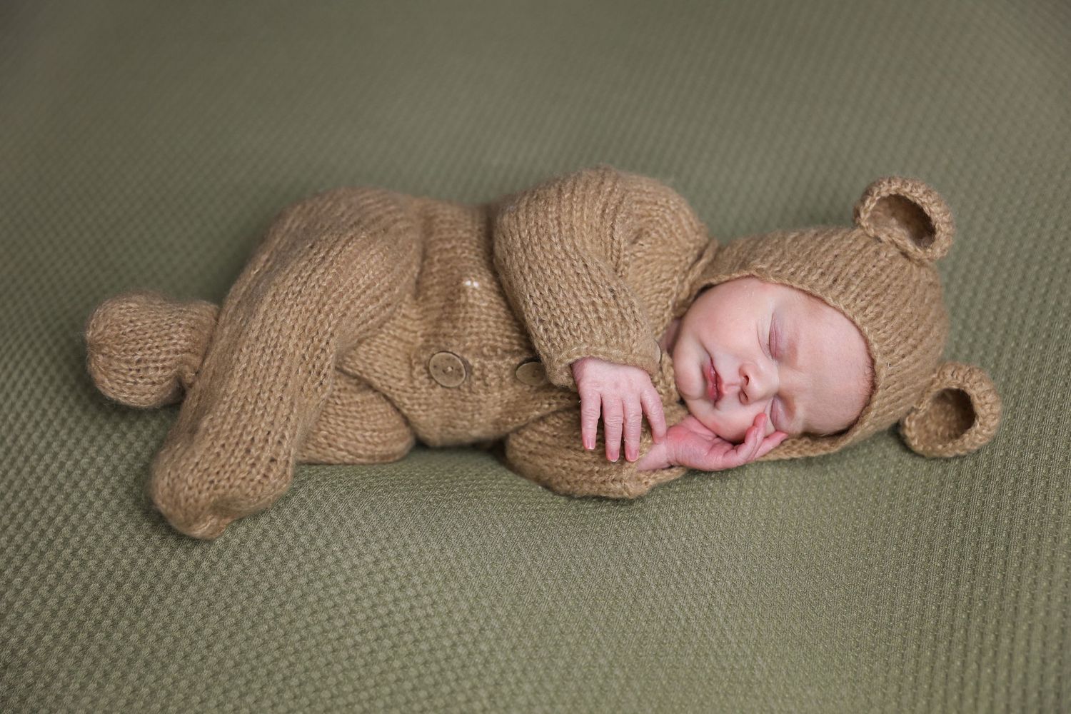 Newborn dressed in knitted teddy bear outfit sleeping on textured green surface.