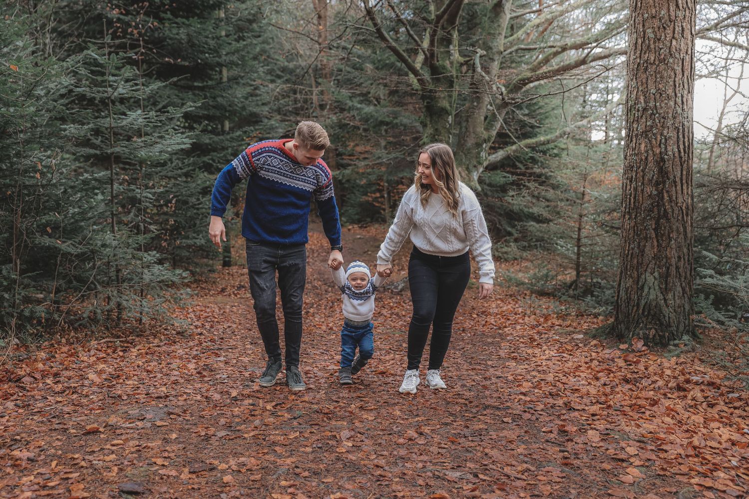 Family walks together holding hands on autumn forest trail covered in fallen orange leaves.