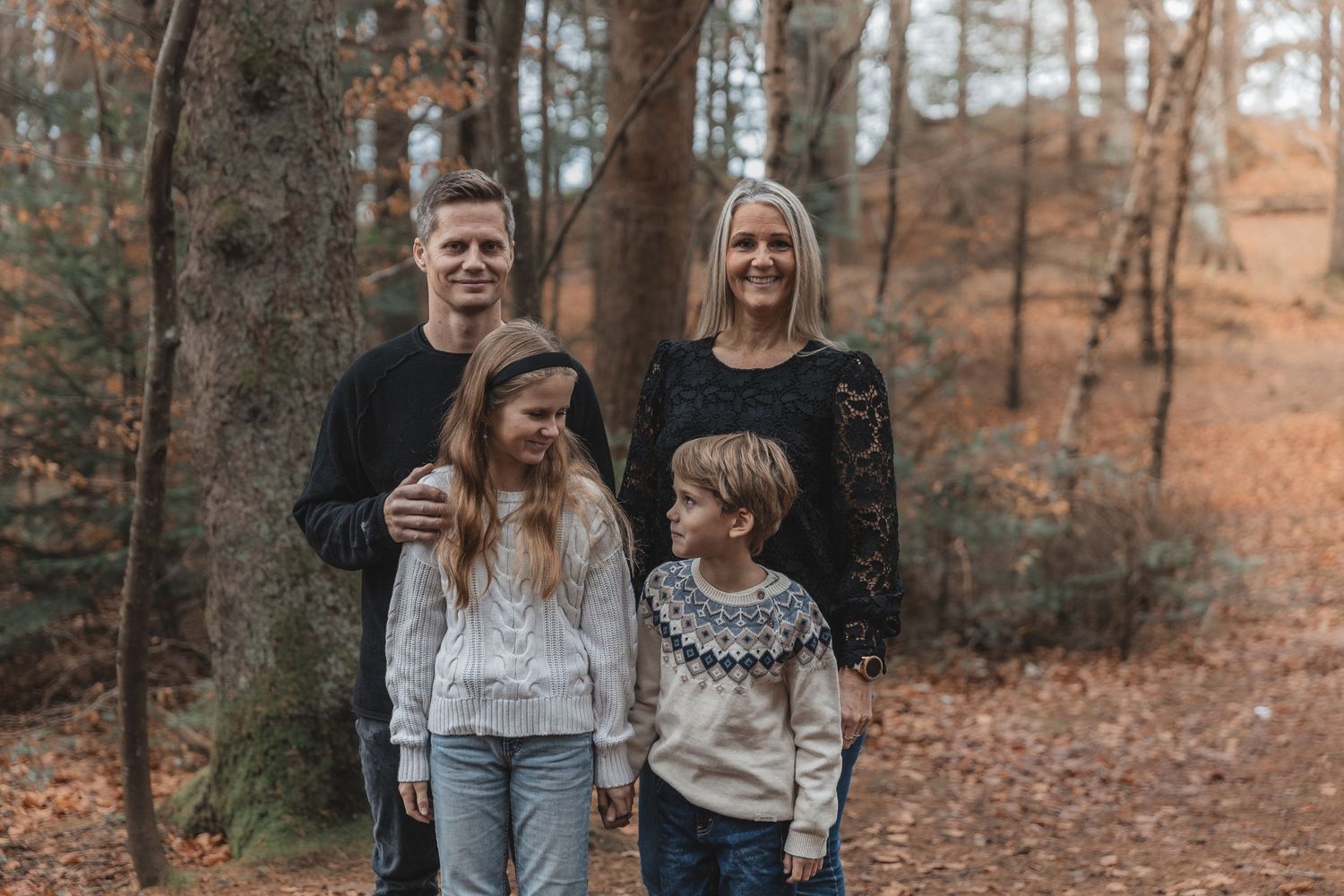 A family smiles together while standing in a wooded area with autumn foliage in the background.