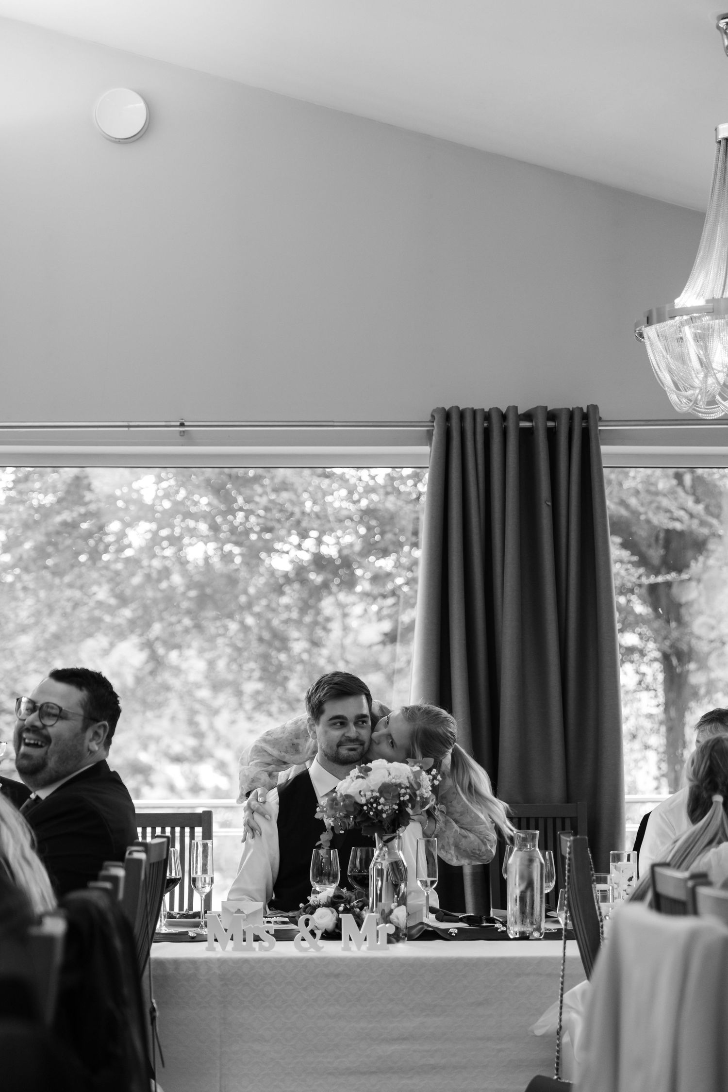 Black and white wedding reception photo of a bride kissing groom at head table while guests laugh and celebrate.
