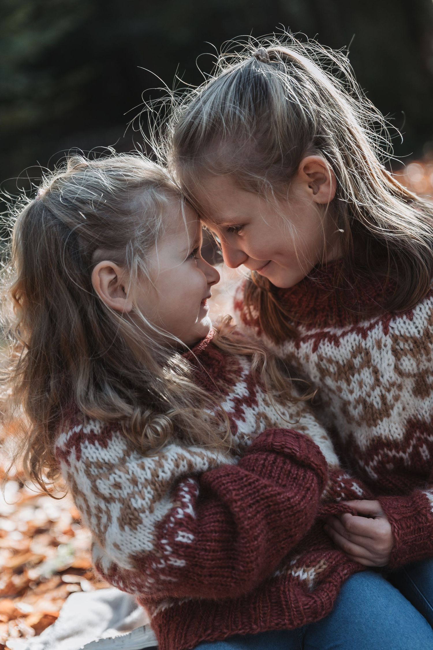 Two young sisters in matching Nordic sweaters touch foreheads affectionately outdoors in autumn.