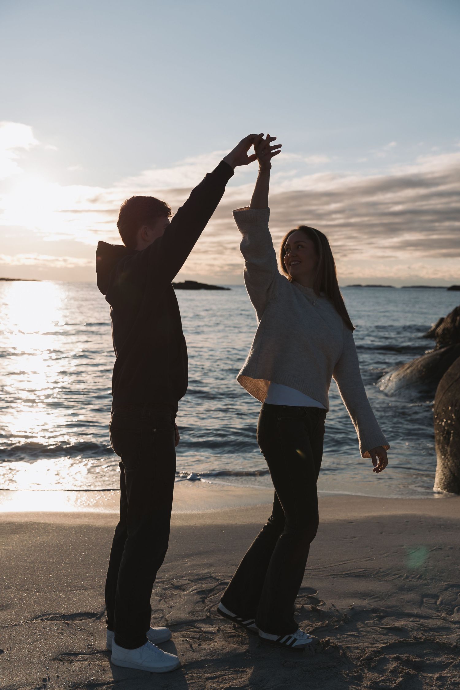 Couple dancing on a beach at sunset, holding hands raised, with ocean waves and golden sky in the background.