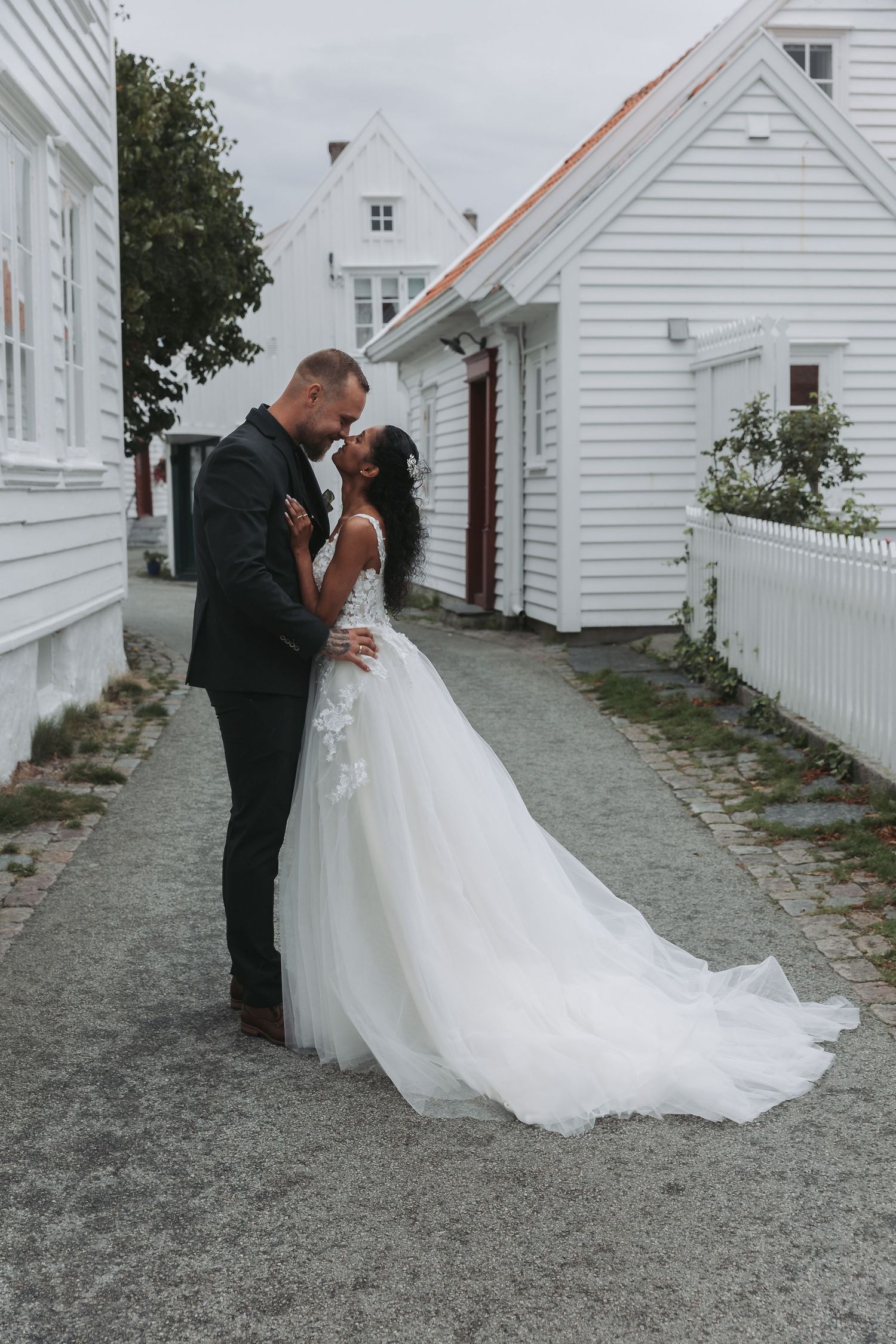 Bride in white tulle gown and groom in black suit share romantic moment between white wooden buildings.