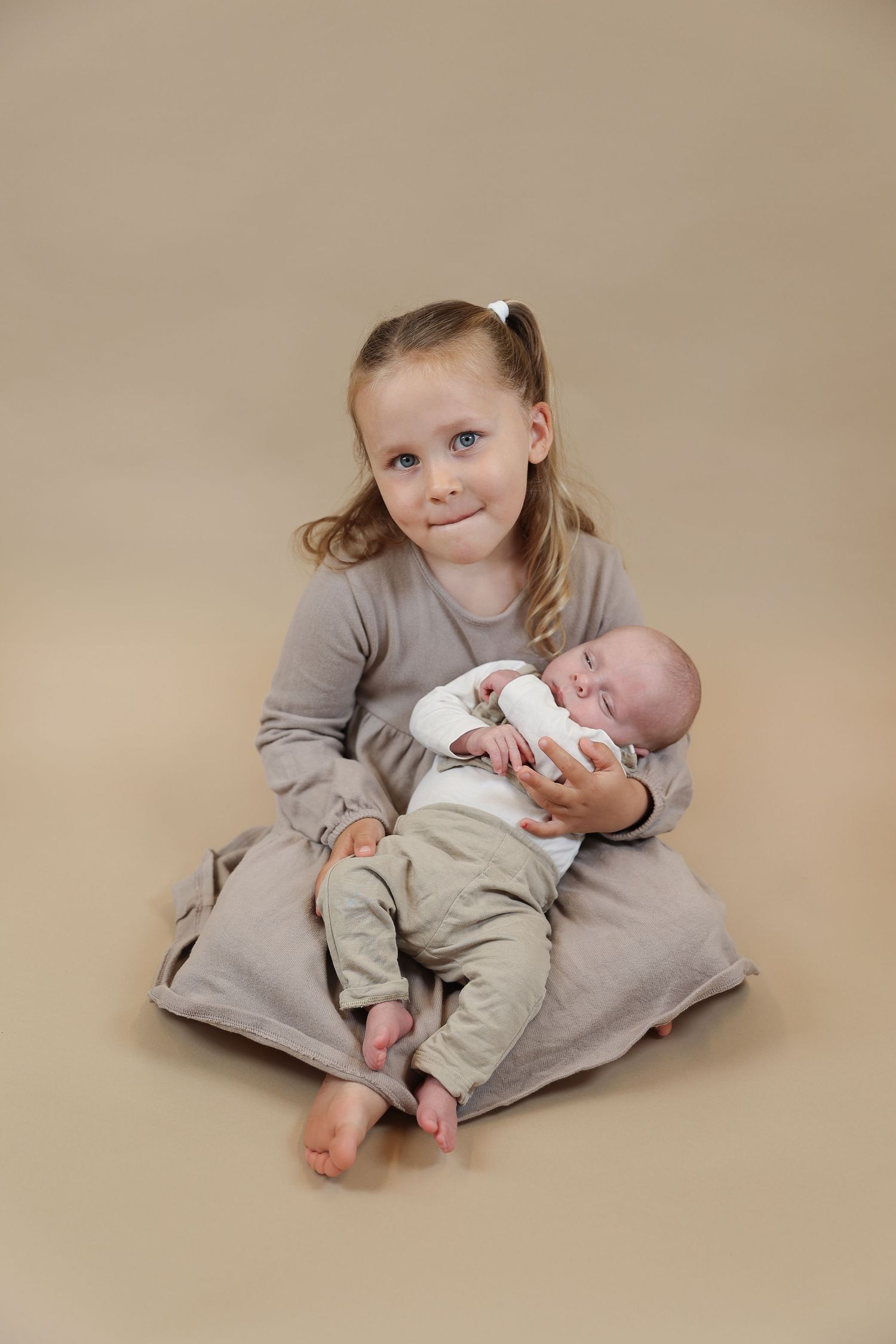 A young child sits on a beige backdrop holding a sleeping infant, both wearing soft neutral colored clothing.