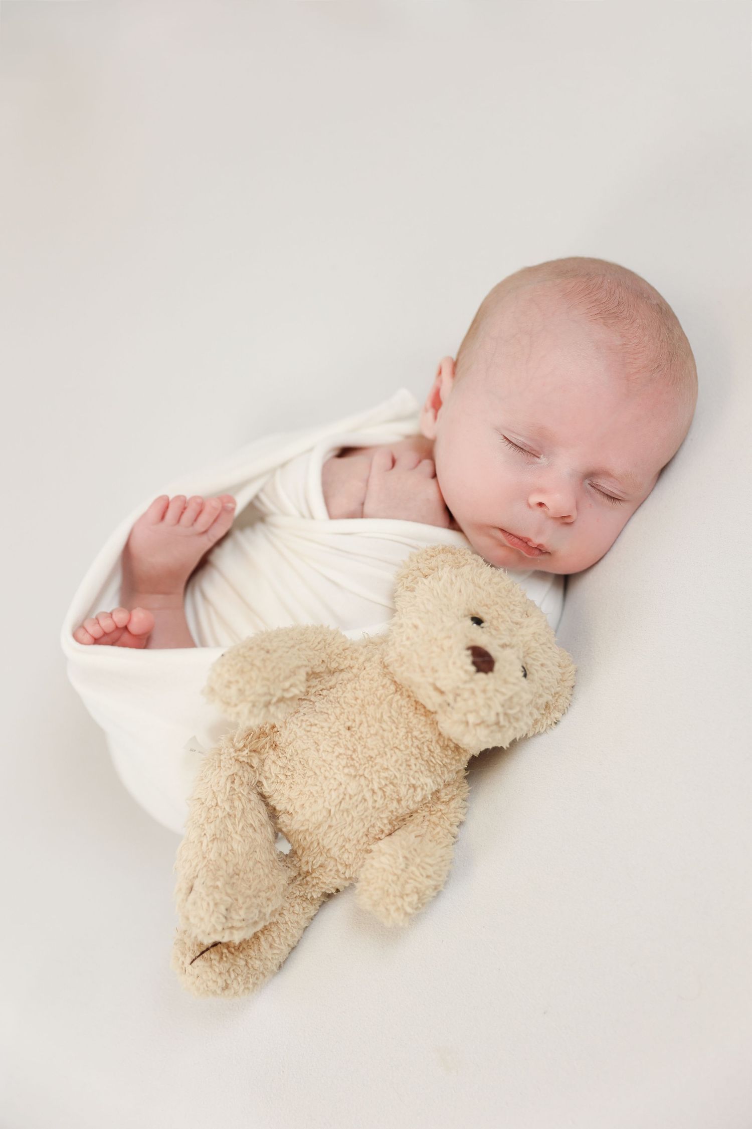 Sweet newborn sleeping in white onesie with plush teddy bear on light backdrop in series of four poses.
