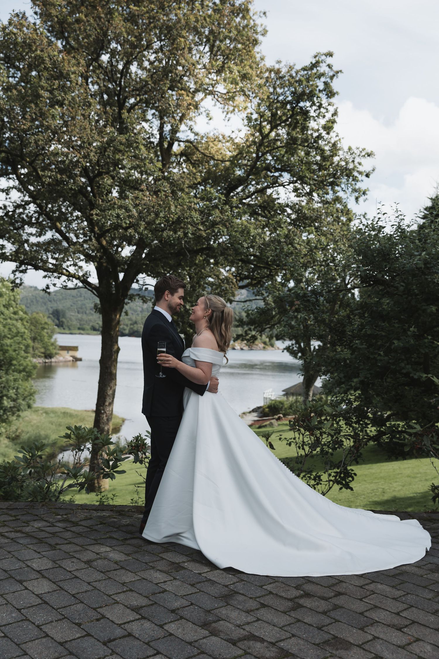 Bride and groom embracing on terrace with scenic lake and trees in background.