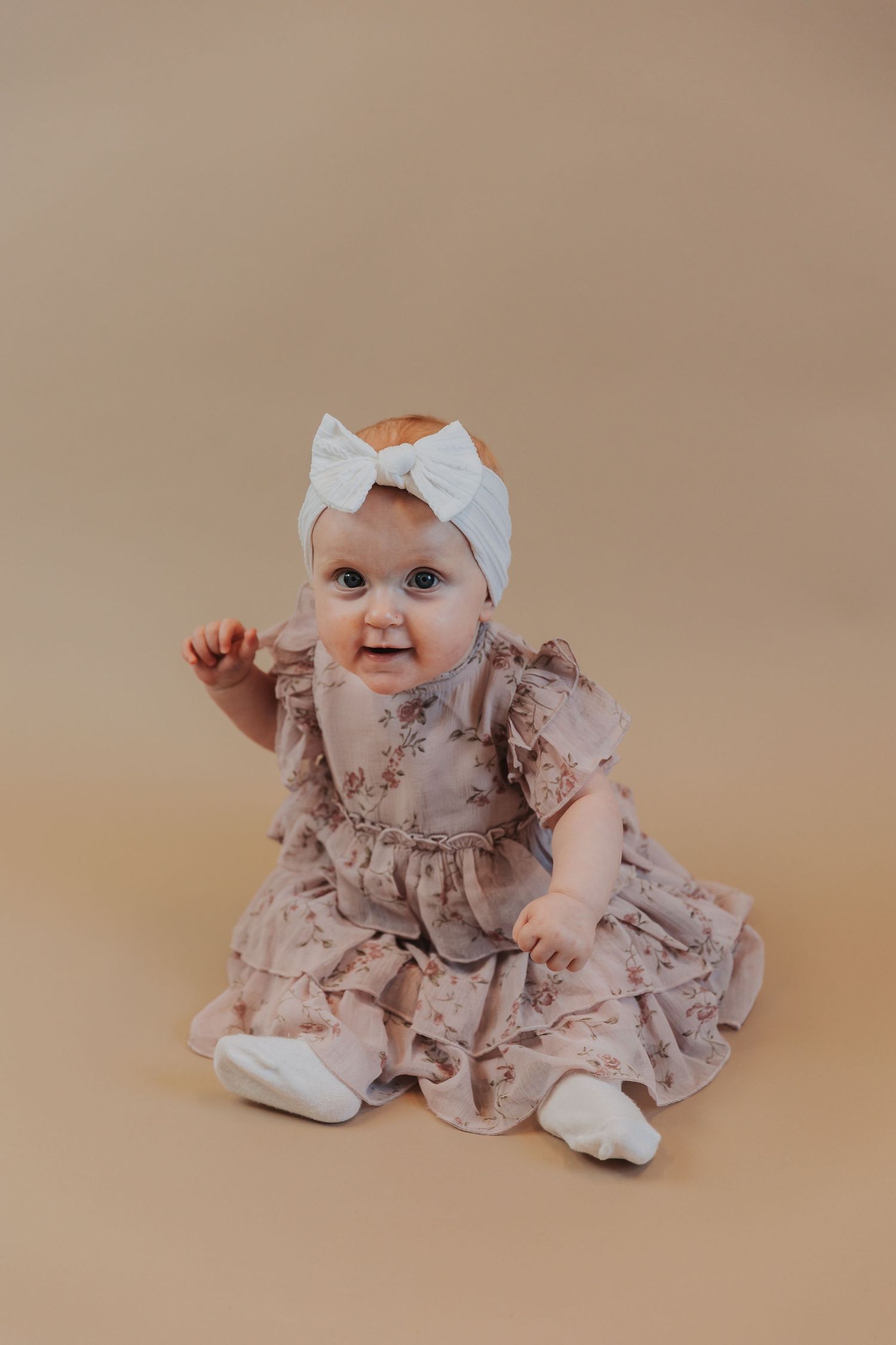 A baby wearing a white headband and dusty pink ruffled dress sits on a beige background.