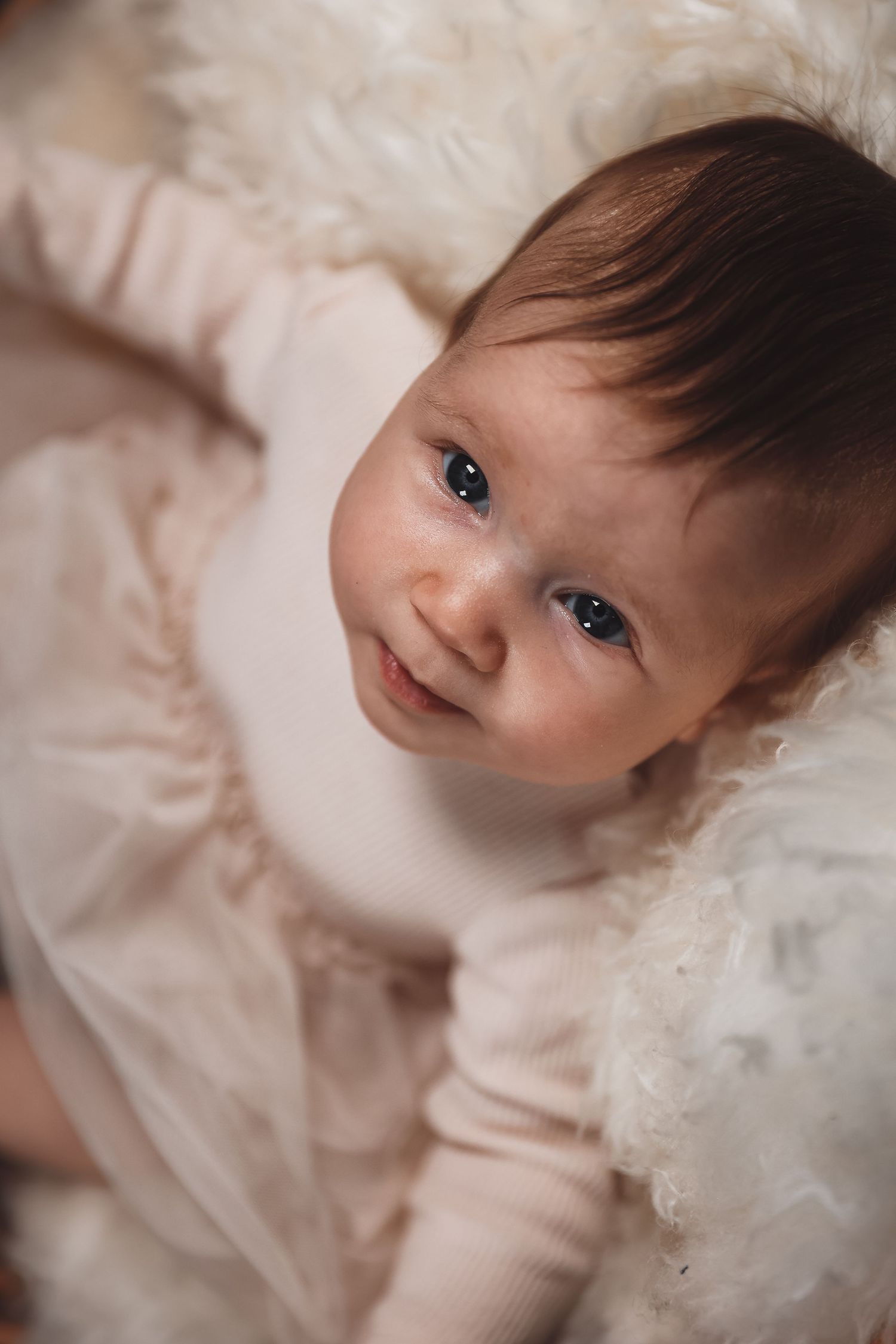 A baby with bright eyes wearing soft pink clothing lies on white bedding.