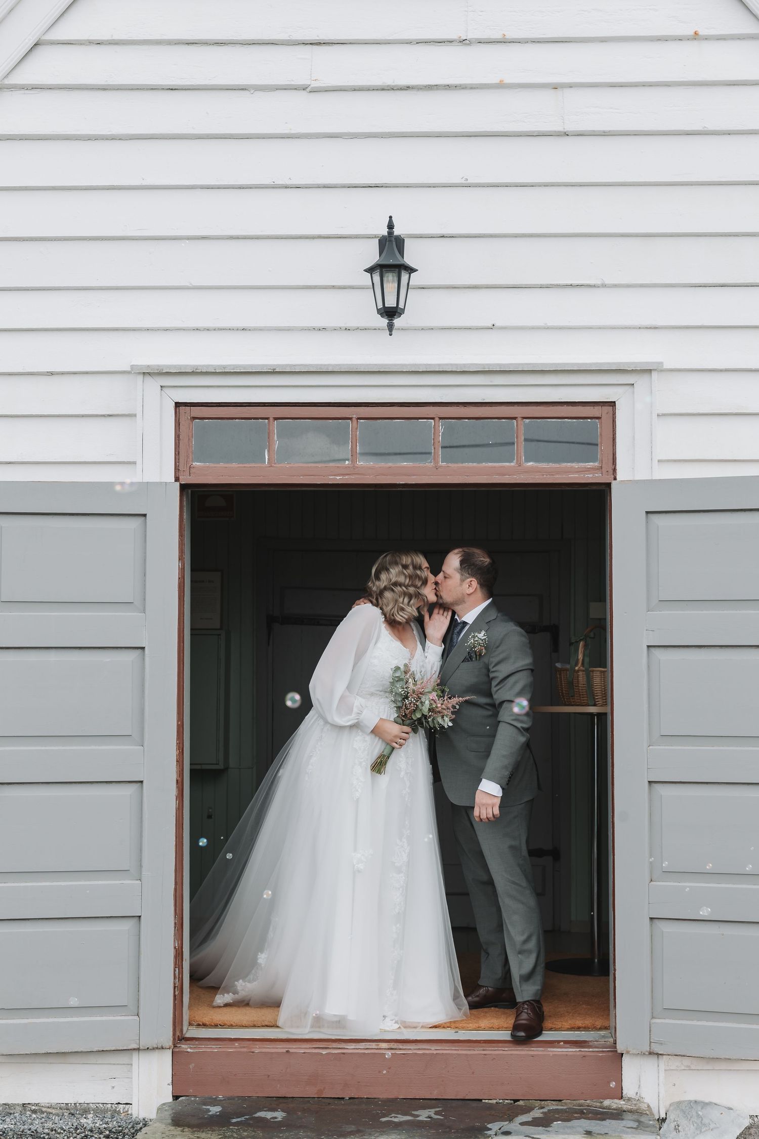 Bride and groom sharing a kiss in a rustic white chapel doorway, bride holding a bouquet in a flowing white gown.
