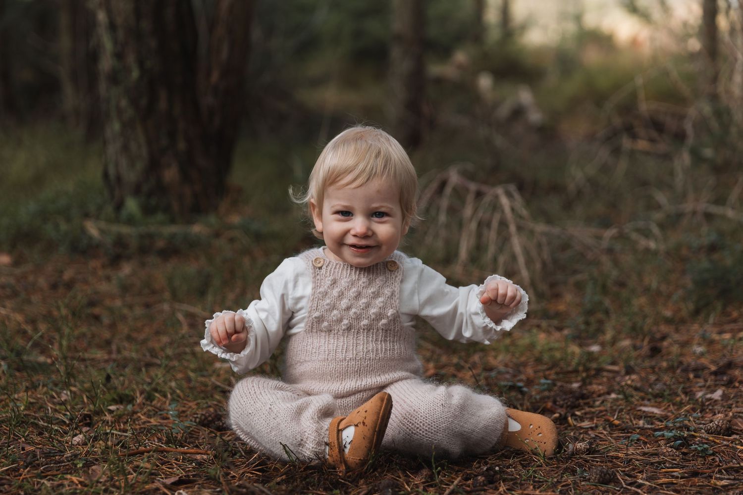 A happy toddler in a cream knit outfit sits on forest ground, smiling with arms outstretched among pine trees.