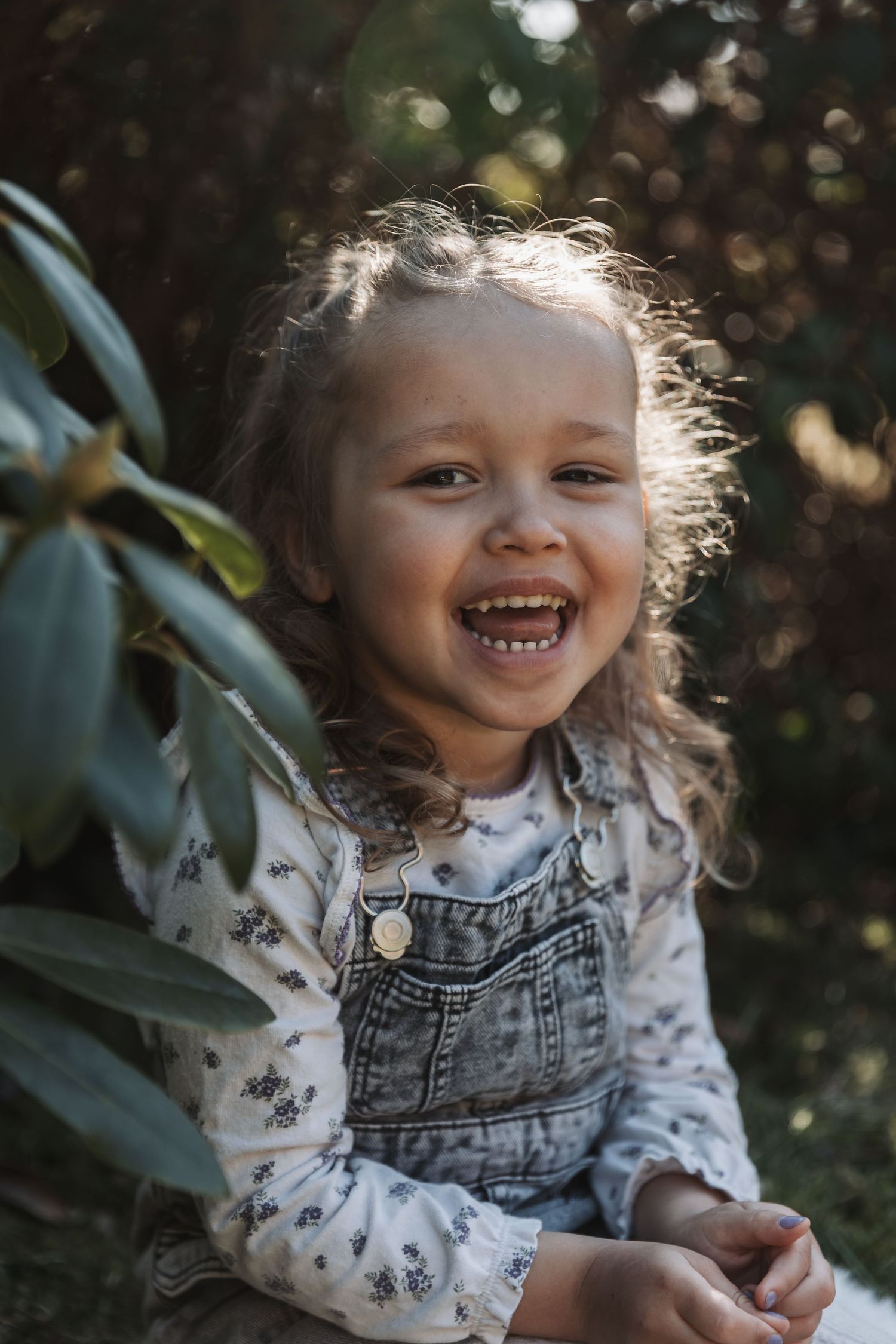 A joyful toddler girl in denim overalls laughing outdoors surrounded by lush green foliage in warm sunlight.