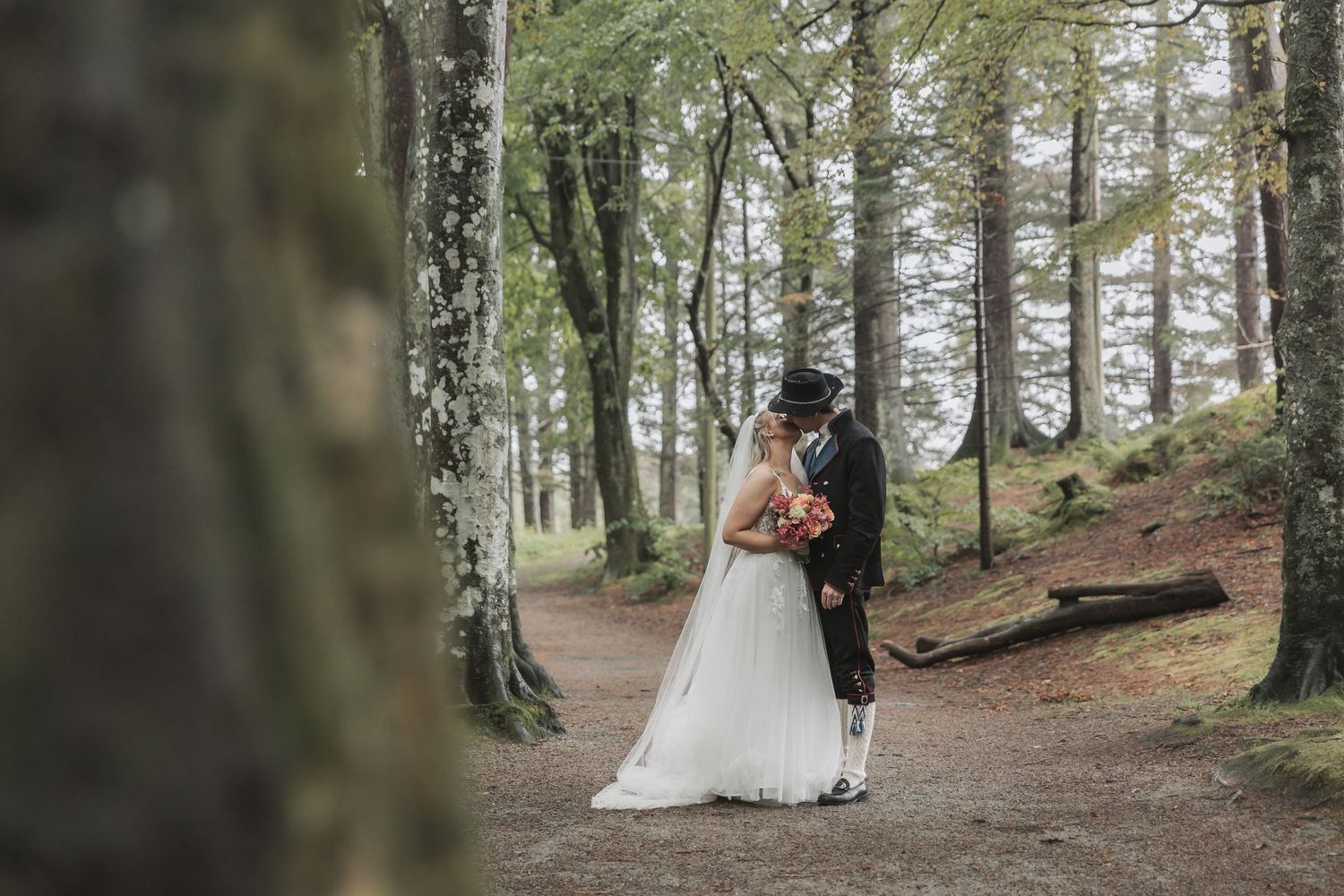 A couple in wedding attire shares a romantic moment in a misty forest setting with tall trees.