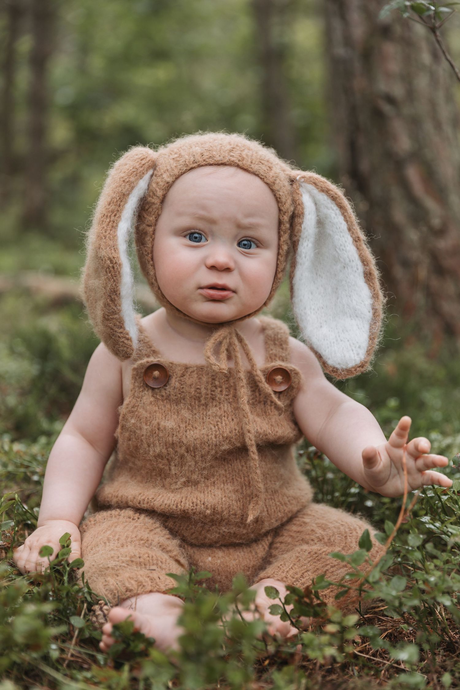 Infant dressed in soft brown bunny costume with long ears sits on forest floor among greenery.