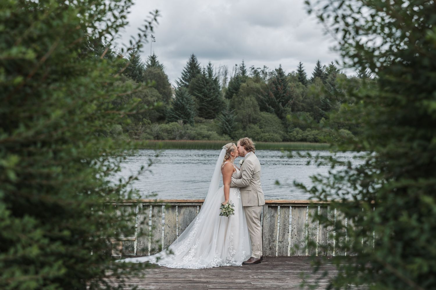 A romantic wedding couple embraces on a lakeside dock surrounded by evergreen trees on an overcast day.