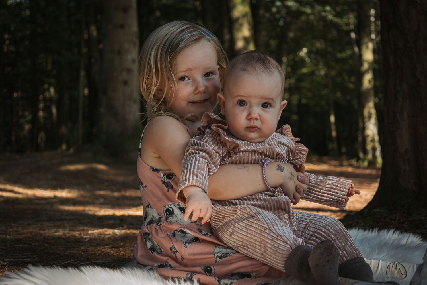 Two children sit together on white fur in a sunlit forest setting.