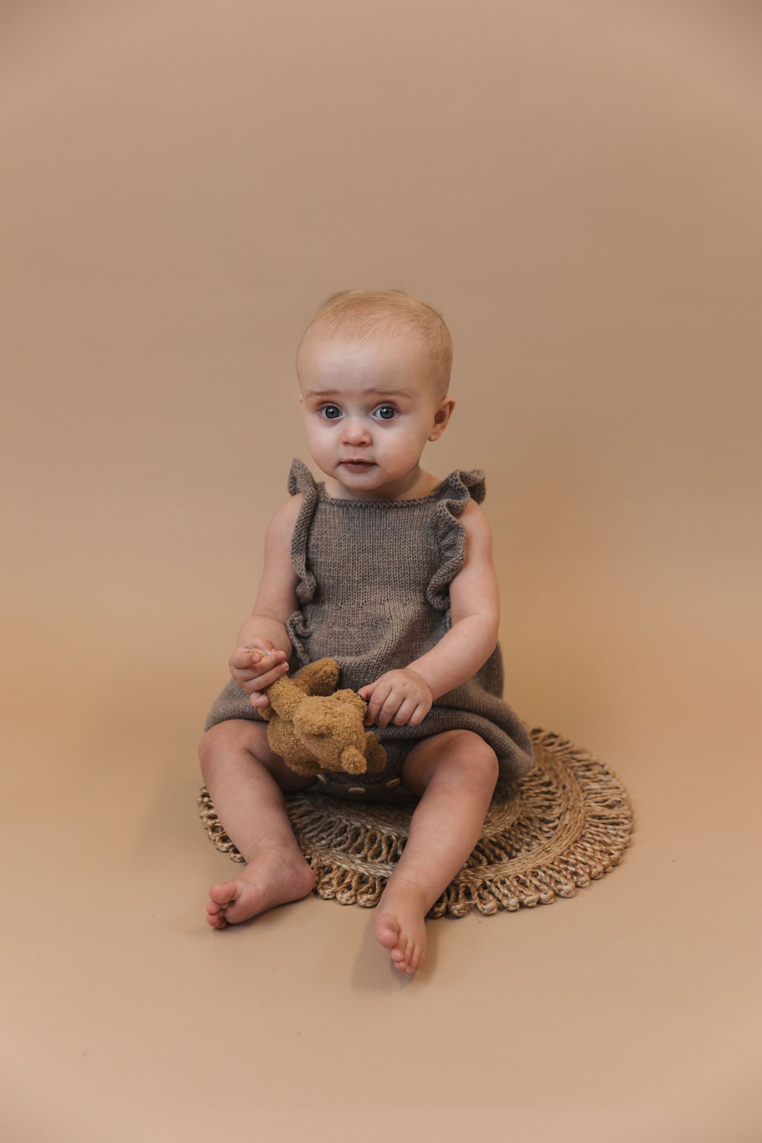 A baby in a gray ruffled romper sits on a woven mat holding a teddy bear against a neutral beige background.