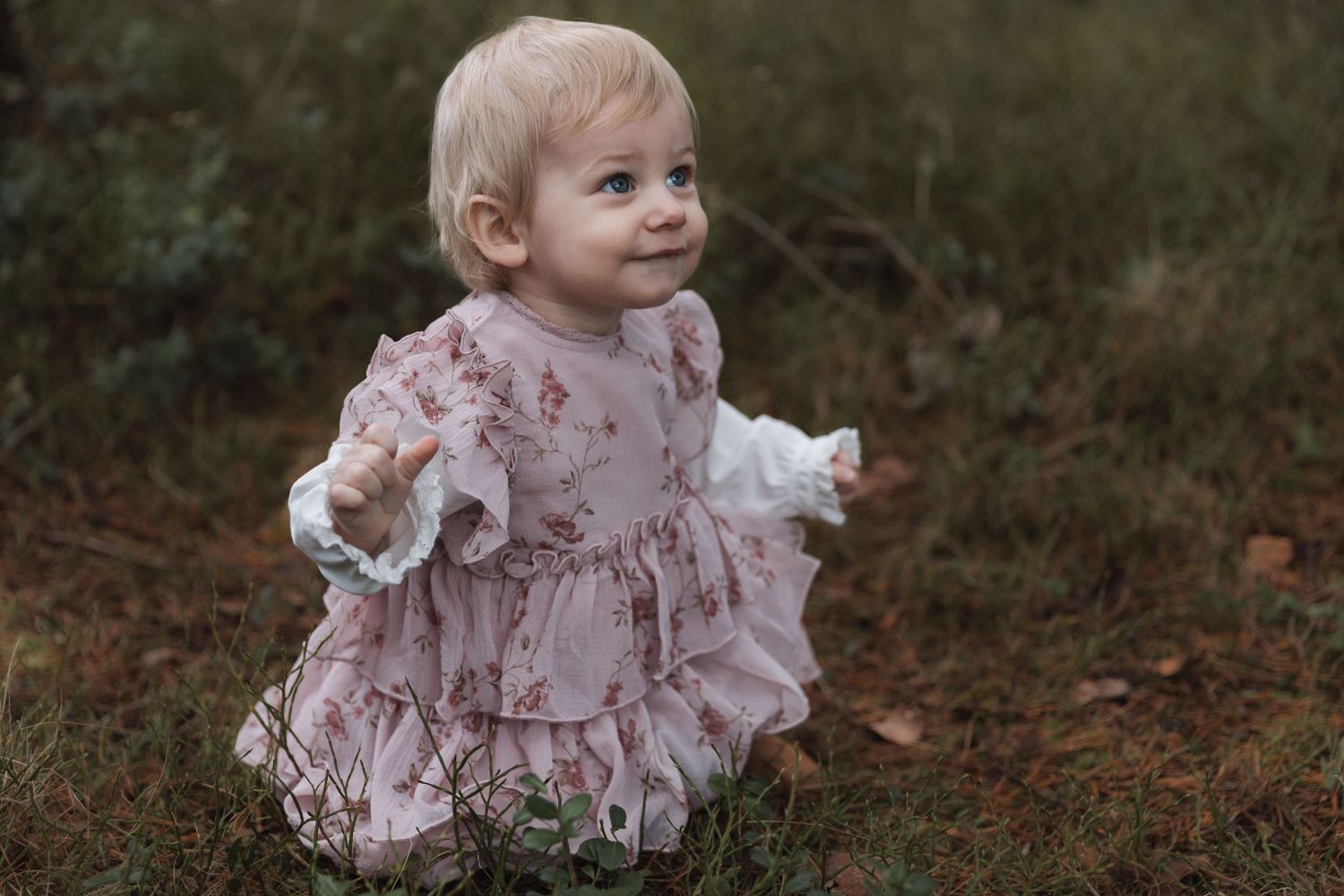 Toddler wearing a pink ruffled dress explores outdoors on an autumn day, looking happy and playful.