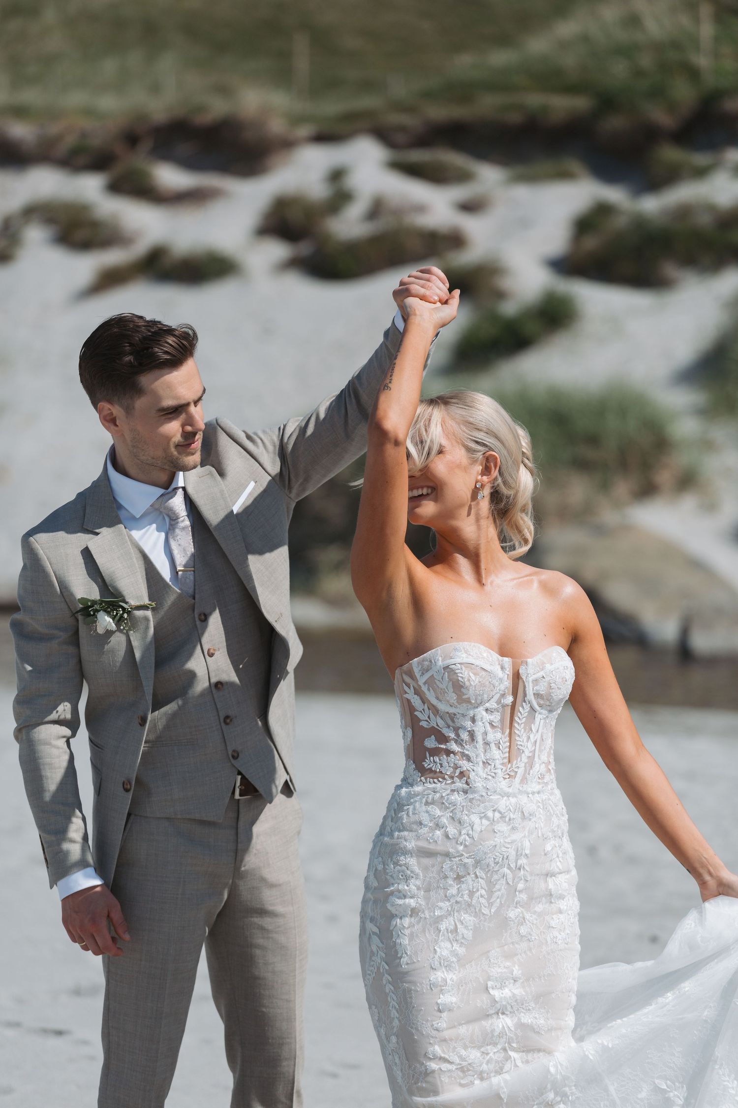 Bride and groom dancing on a sandy beach, she wears a lace strapless gown, he wears a grey suit.
