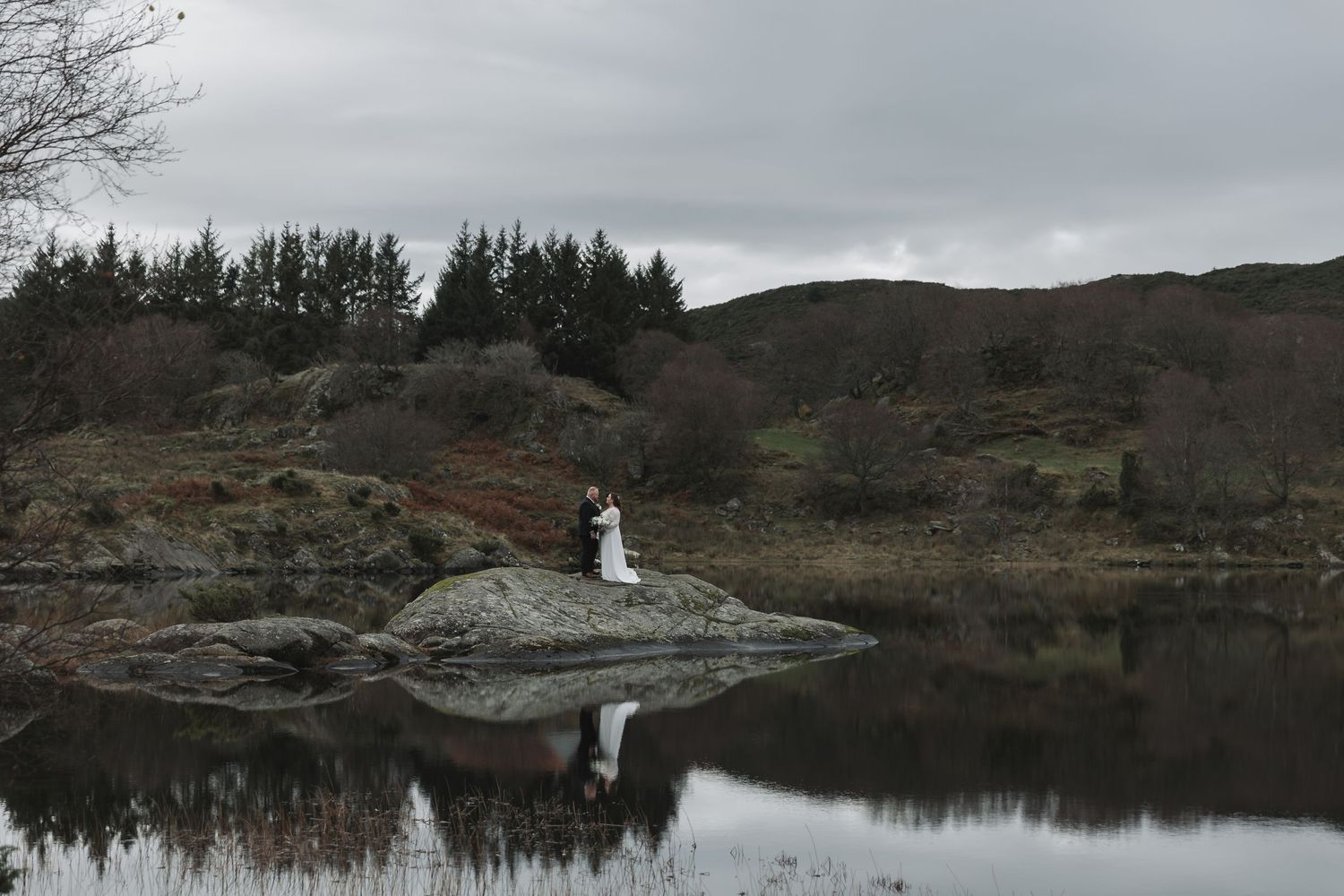 Bride and groom standing on a rock by a reflective lake surrounded by lush hills and trees on a cloudy day.